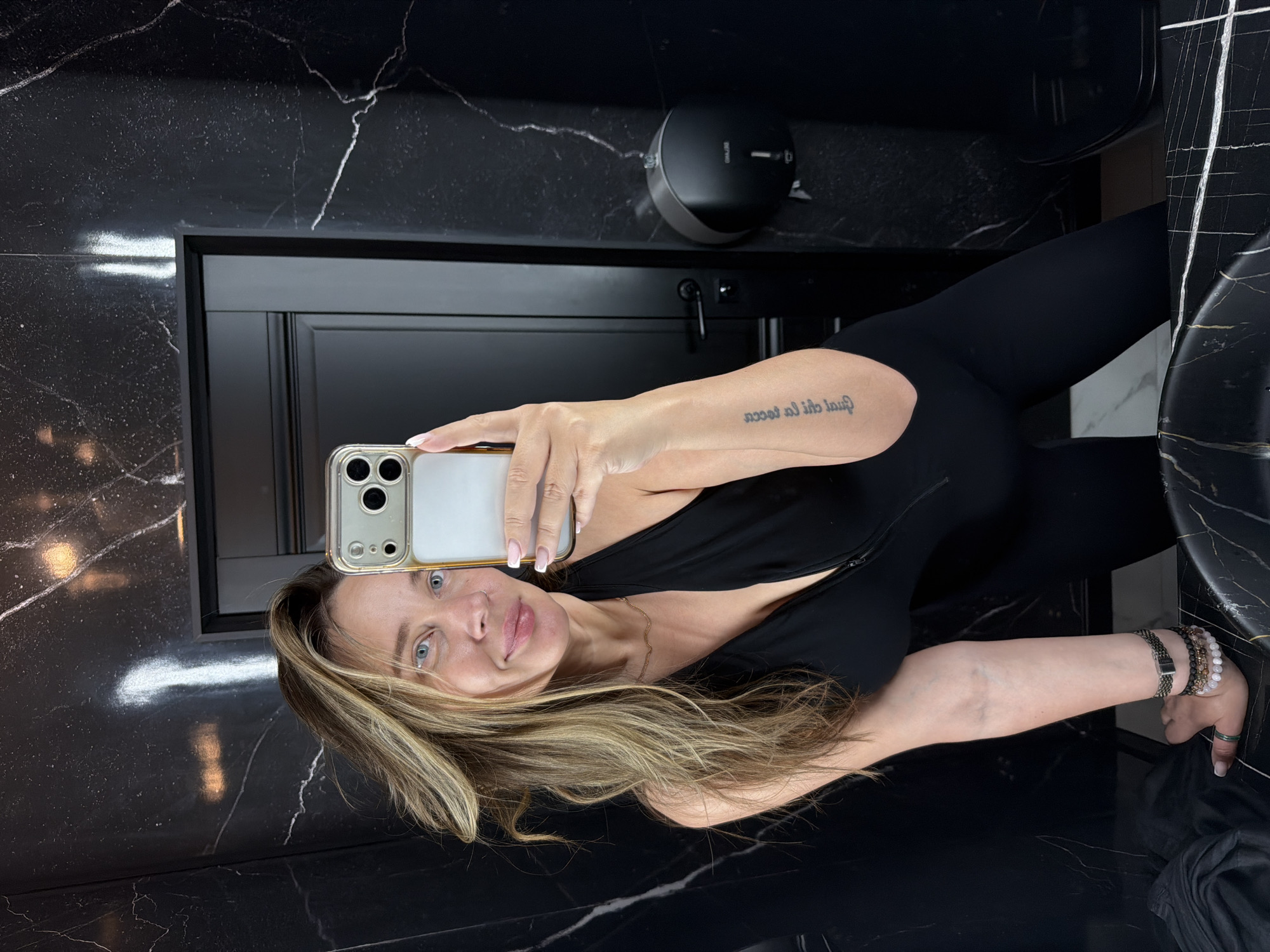 A woman is taking a selfie in a dimly lit bathroom with black marble walls. She is wearing a black, sleeveless jumpsuit with a zippered neckline. She has blonde hair that is slightly windswept, and she is wearing several bracelets on her left wrist. A tattoo is visible on her left forearm. In the background, a black door and a black dispenser are visible. The woman is holding a smartphone in her right hand, which is angled to capture the selfie.