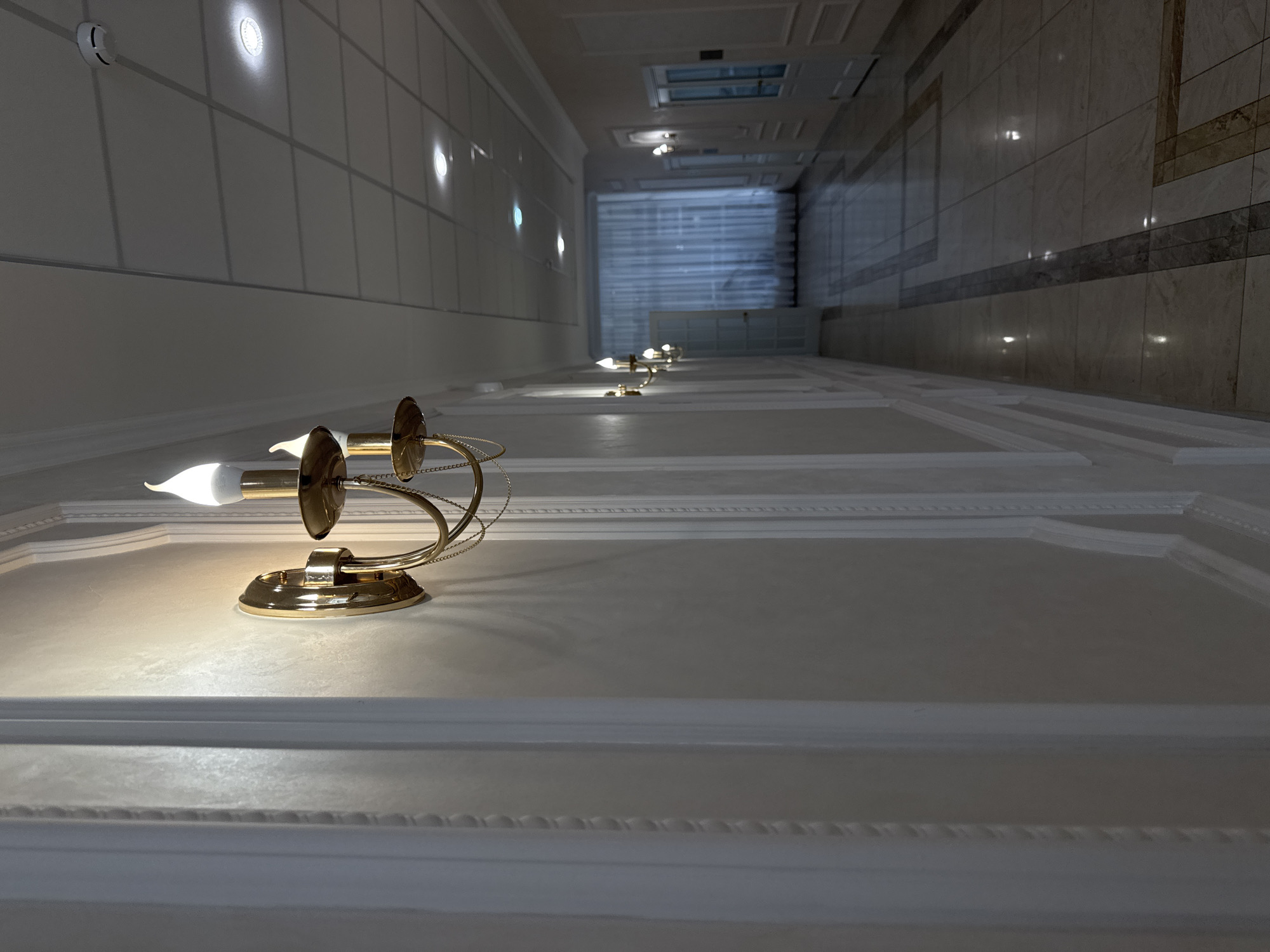 The image captures a long corridor with a series of ornate wall sconces illuminating the space. The corridor is characterized by its perspective view, stretching into the distance towards a brightly lit window at the far end. The walls are adorned with contrasting textures, one side appearing to be a smooth, paneled surface with recessed lighting, while the other exhibits a tile-like pattern. The focus is drawn to the wall sconce in the foreground, which features a gold-colored base and a curved arm holding a small, illuminated bulb. The ceiling has decorative moldings, adding to the overall aesthetic of the space.
