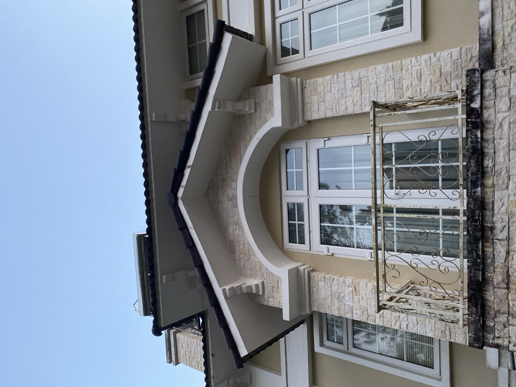 The image shows a portion of a building facade in Da Lat, Vietnam. The architectural style incorporates elements of European design, as evidenced by the arch over one of the windows, the white trim, and the use of light-colored stone. The building has multiple windows, some of which have decorative ironwork. The stone facade appears to be textured, possibly imitation stone or brick. The sky is visible in the background.