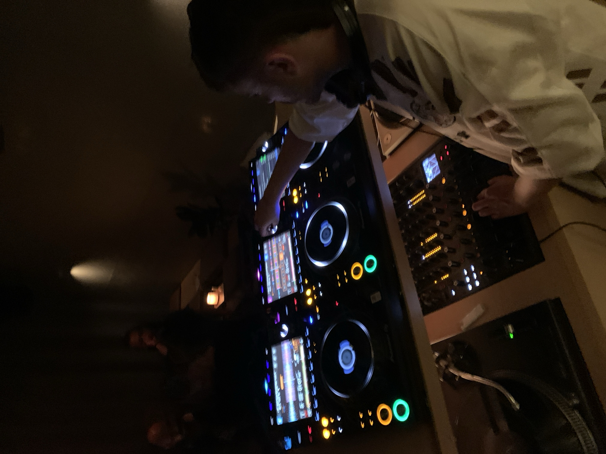 A DJ operates equipment in a dimly lit Tel Aviv club. The DJ, wearing headphones and a white shirt, is angled over the DJ equipment. The equipment includes two CDJ decks with illuminated screens and lit jog wheels, a mixer, and a turntable. The surrounding environment is dark with minimal ambient lighting.
