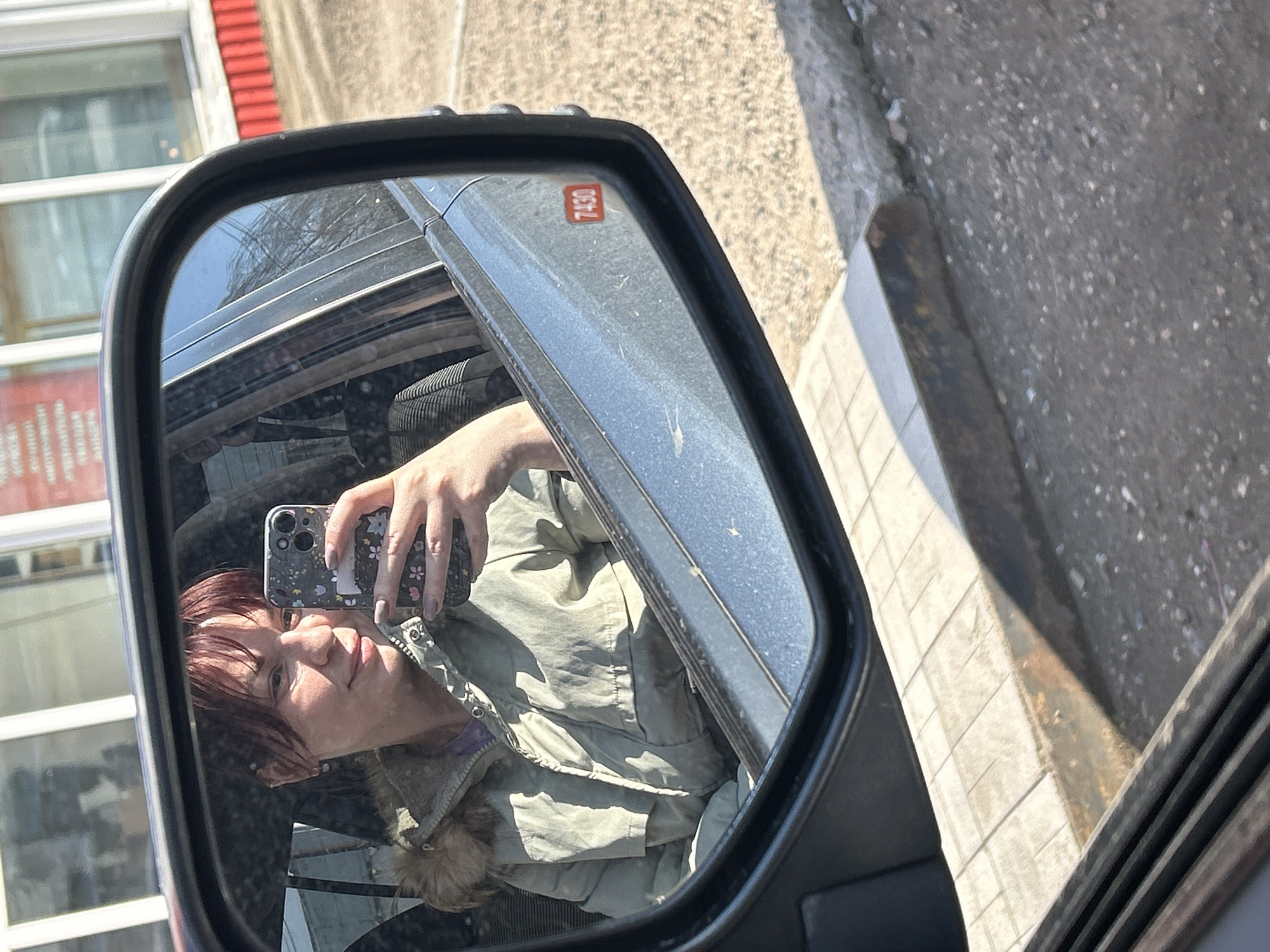 A woman with short red-brown hair is taking a selfie with her cell phone in the driver's side mirror of a vehicle. The mirror is reflecting the interior of the car and her reflection. The background shows a building with a red and white facade. The vehicle has a sticker with the number "7430" on it. The street and sidewalk are visible outside the car. Based on the information provided, this image was taken in Chaikovsky, Russia.