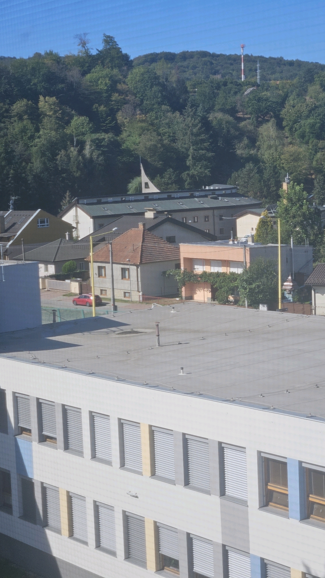 The image depicts an aerial view of a residential and institutional area in Prešov, Slovakia, likely during daytime under a clear blue sky. In the foreground, a modern building with multiple floors and many windows covered by grey blinds dominates the lower portion of the frame. The building's facade is white with vertical grey and yellow accent panels.
In the mid-ground, a cluster of smaller buildings, including houses with pitched roofs, are visible. One house features an orange roof, and another is painted a light orange. A red car is parked on a street or driveway near these houses. A yellow pole, possibly a sports goalpost, stands prominently.
The background is characterized by a densely forested hillside. At the summit of the hill, a telecommunications tower with red and white markings is visible, along with other smaller structures and antennas. The overall scene suggests a suburban or semi-rural setting within the city of Prešov. There are no people or animals visible in the image. The perspective is from a high vantage point, looking down and across the landscape.