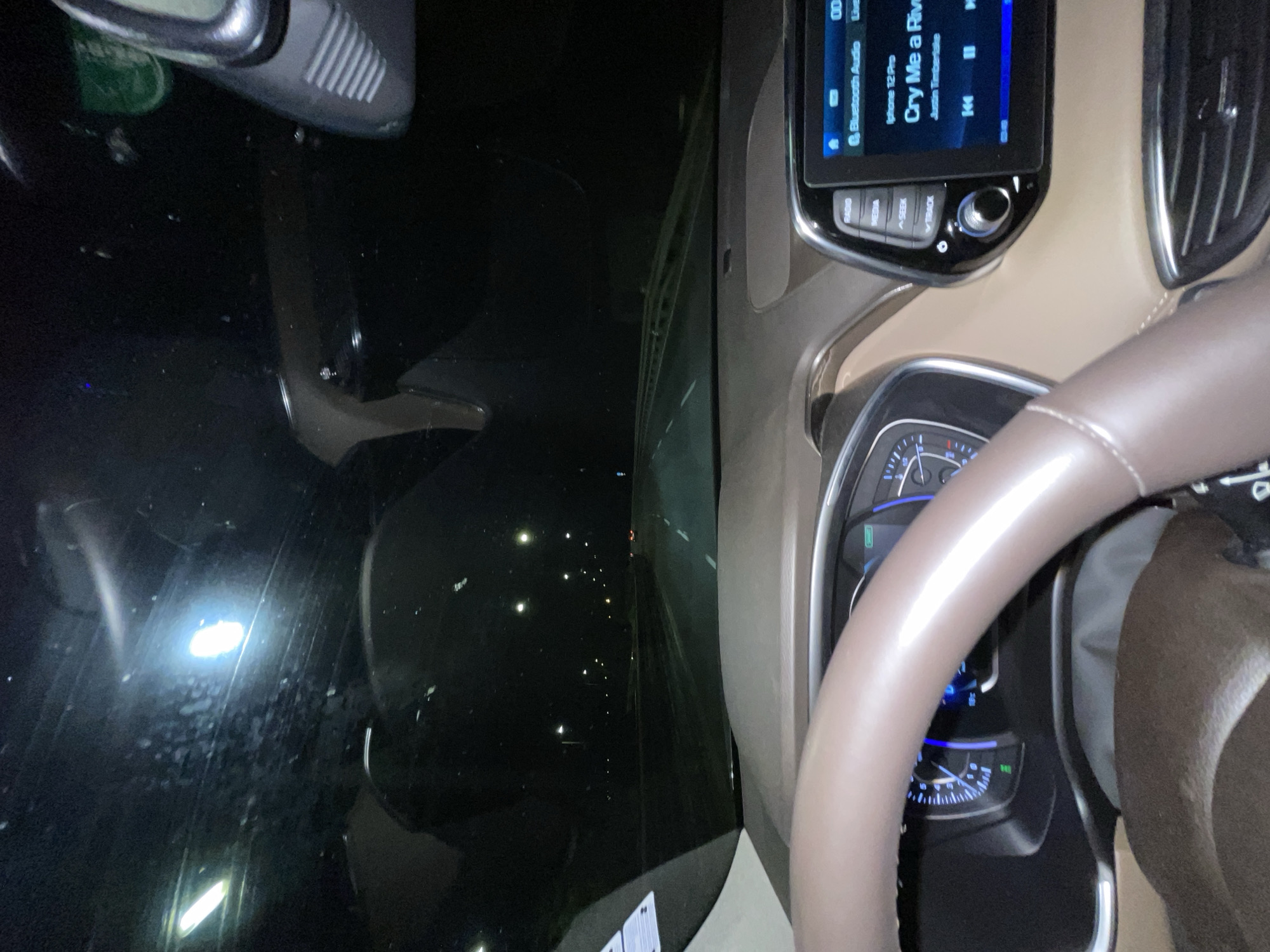 The image shows the interior of a car at night, focusing on the dashboard and steering wheel. The infotainment screen displays "Cry Me a River" by Justin Timberlake. The driver's side window shows streetlights and other cars. The steering wheel is light brown leather. The dashboard is beige. The location appears to be within a city, as indicated by the presence of streetlights and other cars in the distance.