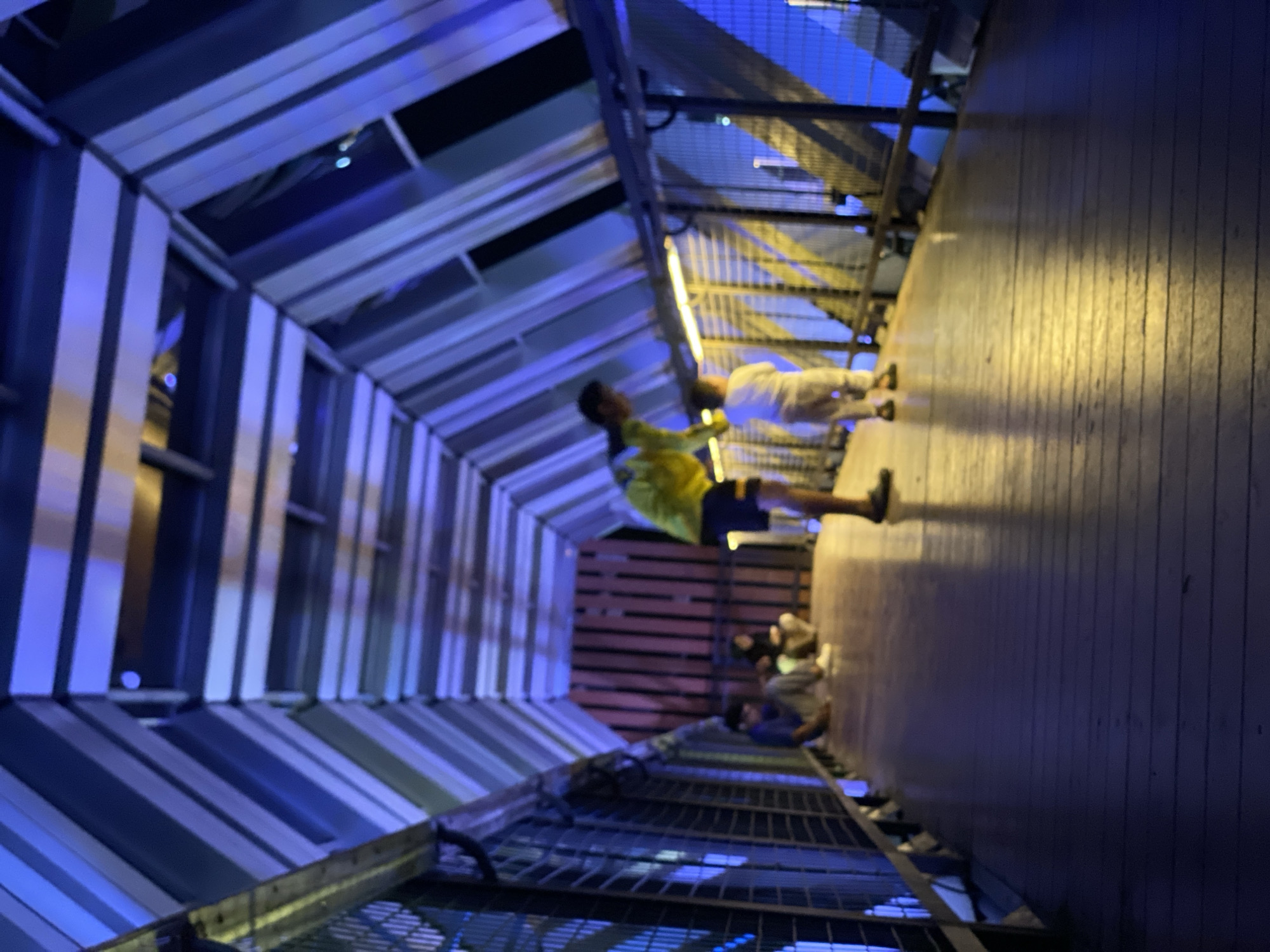 The image captures an indoor pedestrian walkway, possibly an elevated bridge or tunnel, with a unique architectural design featuring angled beams and reflective surfaces that create a geometric pattern overhead. The walkway is illuminated by warm, yellow lights along the sides, contrasting with the cool blue tones of the architectural elements and distant background. Several individuals are present on the walkway. In the foreground, a person wearing a yellow shirt and blue shorts is bending down, seemingly interacting with a child dressed in white. Further down the walkway, two people are seated on the floor, one in a grey shirt and the other in a beige outfit. The floor of the walkway appears to be made of wooden planks. The overall atmosphere is dimly lit and somewhat abstract due to the lighting and the blurriness of the image. The location is identified as Banjarbaru, Indonesia, but no specific landmarks or context within the city are discernible from the image itself.