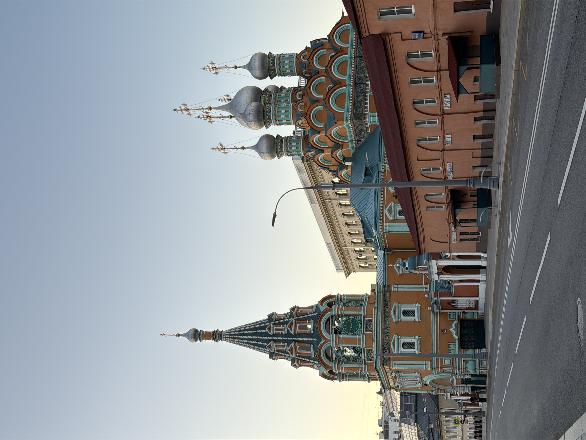 The image depicts the Church of the Holy Trinity in Nikitniki, Moscow, Russia, captured from across a street. The church's distinctive orange facade is adorned with turquoise detailing and features multiple onion domes topped with crosses. To the left, a tall, slender bell tower with a conical roof and similar decorative elements stands prominently. In front of the church, a low, rust-colored building with arched windows and a red roof runs along the street. The street itself is asphalt with white lane markings. A single streetlight stands on the sidewalk. The sky is a clear, pale blue, suggesting it is either early morning or late evening. No people are clearly visible in the immediate foreground or interacting with the buildings. The scene is static and architectural, emphasizing the historical religious structures.