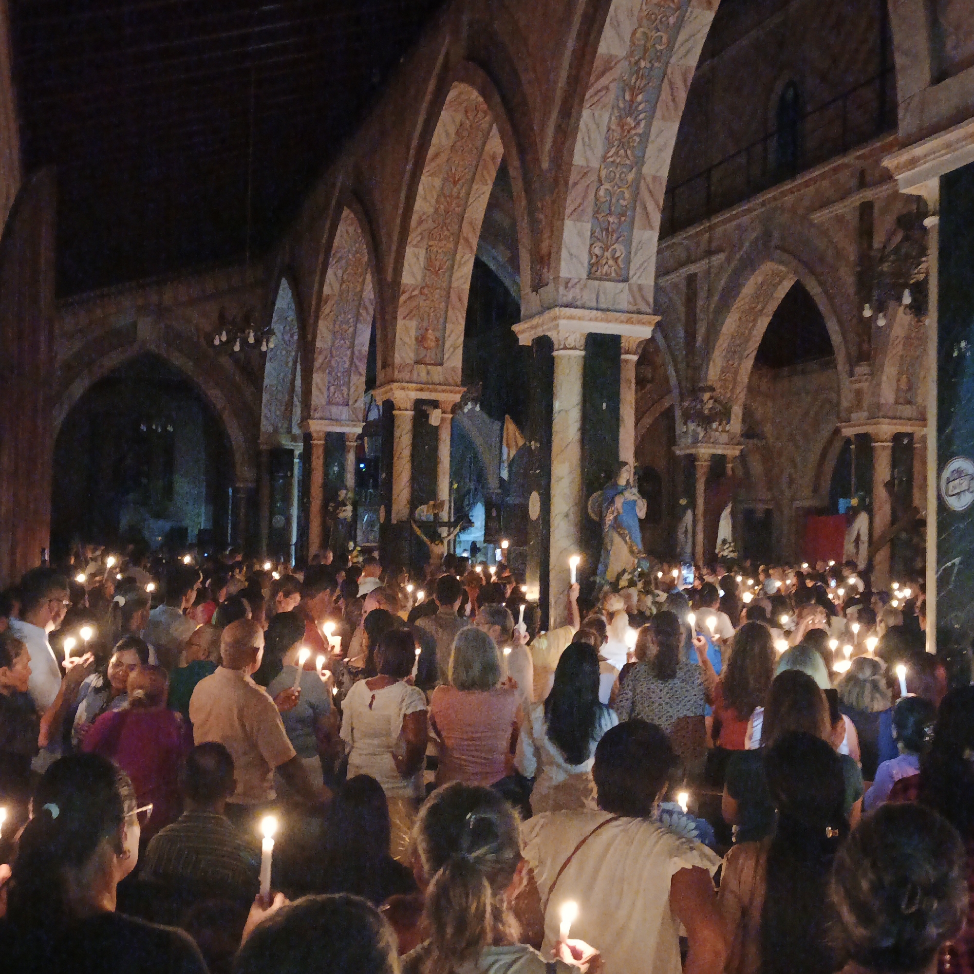 A dimly lit interior view of a church, crowded with people holding lighted candles. The architectural style features arches decorated with floral patterns. The church contains statues and columns. People are primarily facing forward. Location is likely the city of Tinaquillo, Venezuela.