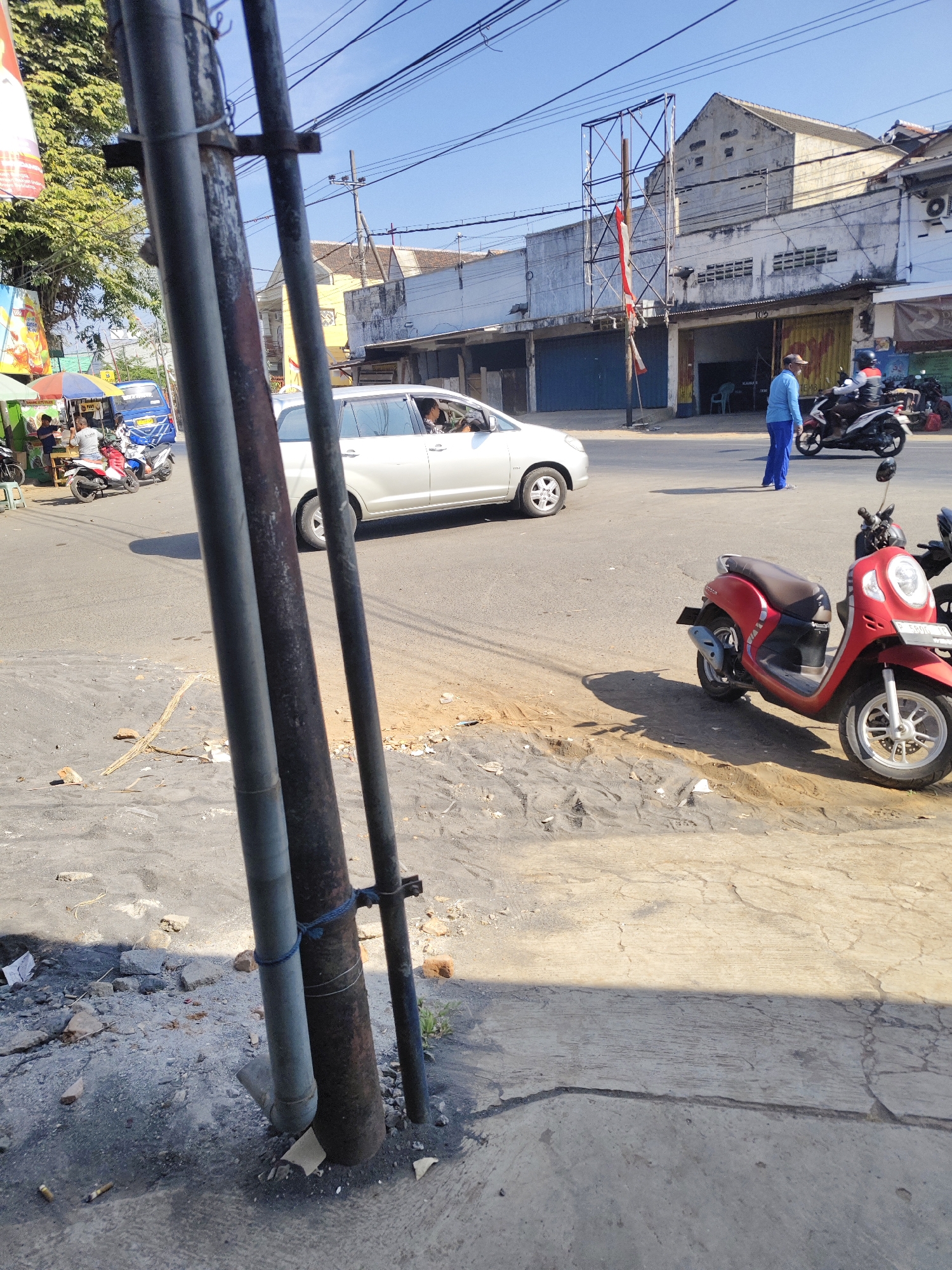 In Gambirono, Indonesia, a street scene unfolds under a clear blue sky. Three tall metal poles stand prominently in the foreground, casting long shadows on the cracked, dusty asphalt. A white van is captured mid-turn, its driver visible through the window. To the right, a red scooter is parked on the side of the road, its license plate partially obscured. Further down the street, a person in a blue uniform walks away from the camera, while another individual on a motorcycle moves past them. The background features a row of buildings with closed storefronts, some adorned with flags and banners. A few smaller stalls with umbrellas are also visible, suggesting a commercial area. The overall impression is a snapshot of daily life on a sunny day in an Indonesian town.