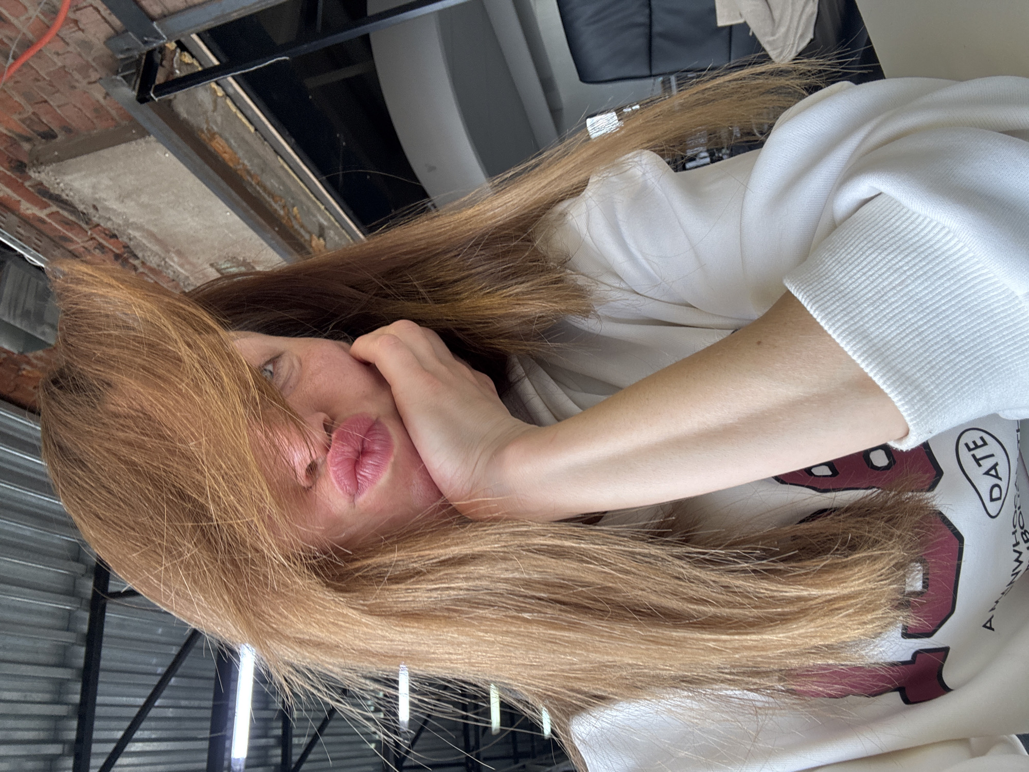 A woman with long, light brown hair is resting her head on her hand. She is wearing a white sweatshirt with the word "DATE" printed on it. The background includes a brick wall, metal structures, and what appears to be a ceiling with various fixtures. The specific location in the city of Chaikovsky, Russia, cannot be determined from the image.