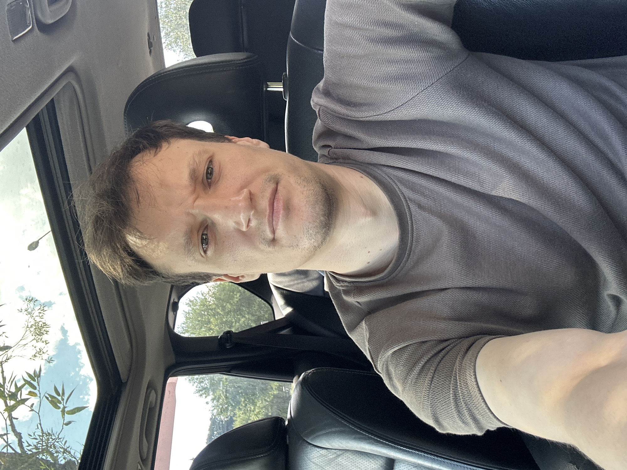 A man is taking a selfie inside a car. He is wearing a grey t-shirt and has brown hair. The car has black leather seats and a sunroof, through which trees and a cloudy sky are visible. The setting appears to be outdoors, during the daytime. The image does not provide specific information to confirm the location as Makiv, Ukraine.