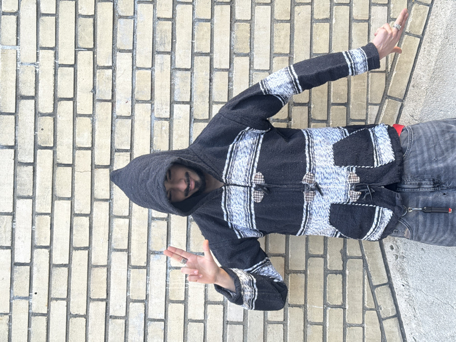 A young individual, appearing to be male, is captured in a full-body shot, standing on a light-colored brick surface. The image is oriented sideways, making the person appear to be lying down against a wall, but in a standard orientation, he would be standing upright on a paved ground.
The person is wearing a dark, textured hoodie with the hood pulled up, partially obscuring the top of his head but leaving his face visible. He has a dark beard or stubble and a subtle smile, suggesting a pleasant expression. His eyes are dark and look towards the viewer.
His hoodie is predominantly black or very dark grey, featuring distinctive horizontal bands of light blue, grey, and white patterns across the chest, back, and sleeves. The fabric has a thick, woven, or knitted texture. It has a central zipper, which is partially open, and notable toggle closures made of what appears to be wood or horn, secured with dark loops, adding to its rustic aesthetic. Two pockets are visible on the lower front of the hoodie, blending with the patterned design.
He is wearing dark grey or black denim jeans. A small hint of a red garment or accessory is visible at his waistline. On his right side (viewer's left), a metal keychain with a small black lighter attached hangs from a belt loop.
Both of his hands are raised in front of him, gesturing. On his right hand (viewer's left), he displays a "peace" or "victory" sign with his index and middle fingers extended, and a silver-colored ring is visible on his middle finger. His left hand (viewer's right) is also raised, with two fingers extended in a similar gesture, and another silver-colored ring is on its middle finger.
The background consists entirely of a pale yellow, beige, or off-white brick pattern, laid in a stretcher bond arrangement. The bricks are rectangular, and the grout lines are visible. This brick surface serves as the ground or floor on which the person is standing. The lighting is bright and even, indicating an outdoor setting during daylight hours.
