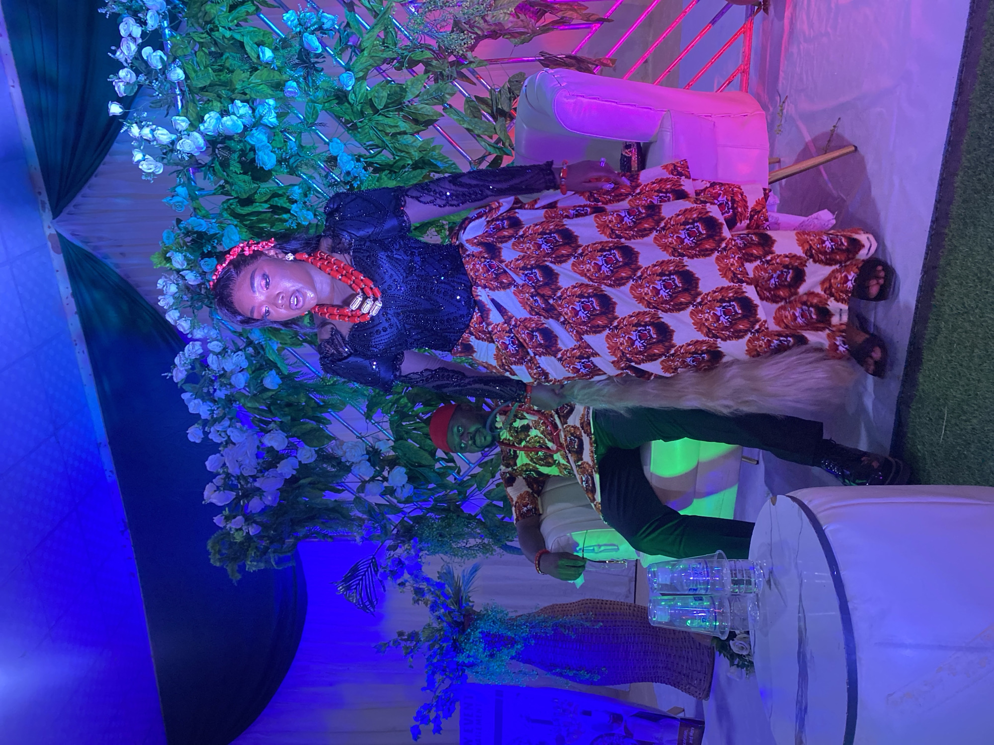 The image depicts a woman in an ornate dress and coral necklace, standing amidst floral decorations. Another individual stands beside her. The setting appears to be a decorated venue, possibly a wedding or celebration. There are tables, chairs, and decorative lighting present. The location is given as Vom, Nigeria.