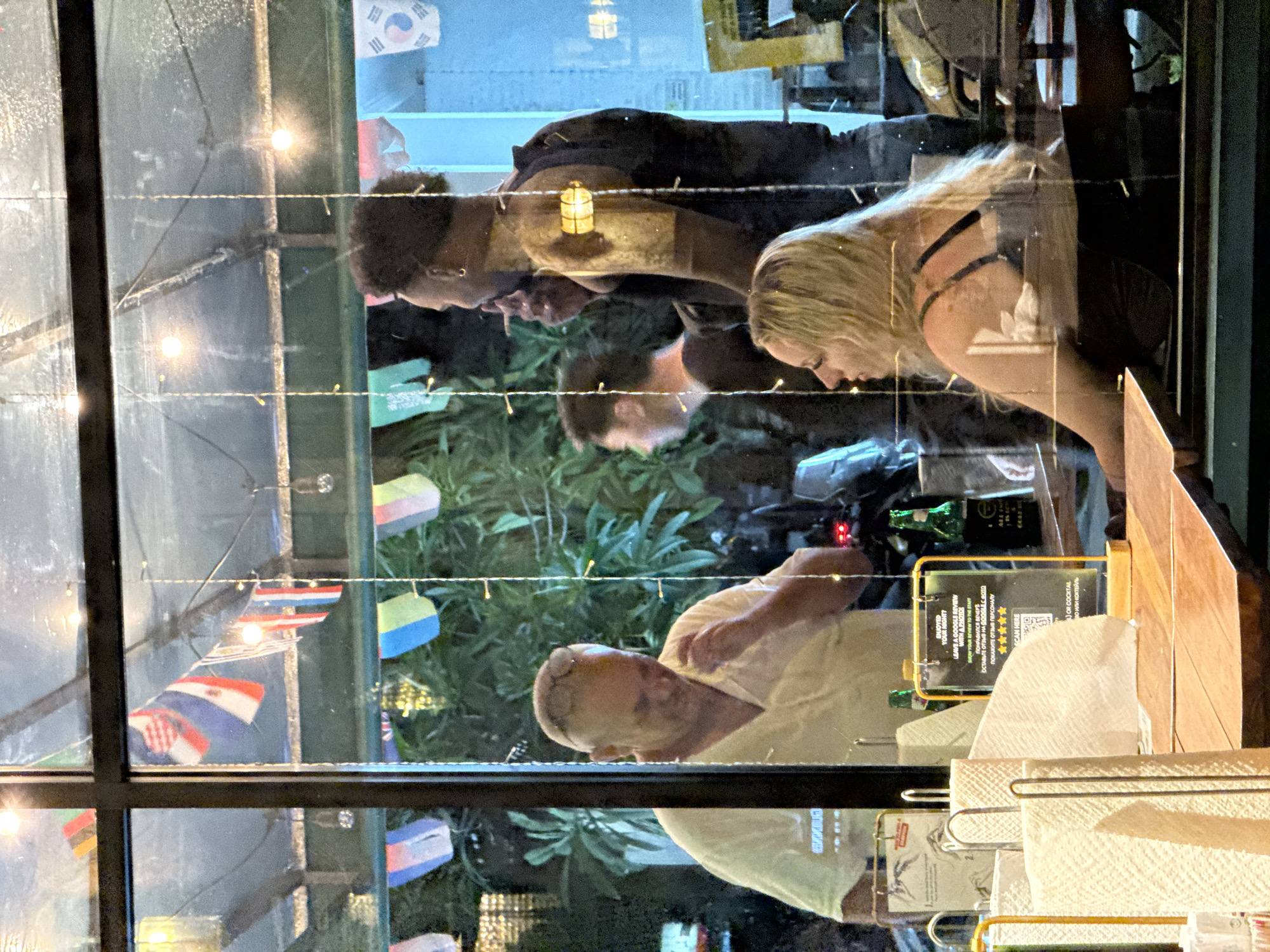 This image captures a scene through a window, likely in Kuta, Indonesia, at night. The reflections on the glass overlay images of people inside with the view of the exterior.
Inside, visible as reflections, are at least four individuals. In the foreground, an older bald man wearing a light shirt and a blonde woman with long hair wearing a dark, strappy top are leaning forward, seemingly looking down at a table. Behind them, two other individuals are reflected, one with dark curly hair and another whose head is partially visible. The reflections suggest they are inside a restaurant or bar, given the presence of a wooden table surface, napkins in a holder, and a small illuminated lantern. A green bottle is also visible on the table.
Outside the window, through the glass, a lush green foliage is visible, illuminated by string lights with small bulbs. Numerous small flags from various countries, including Thailand, Croatia, and South Korea, are strung up, suggesting an international or tourist-friendly setting. The artificial lighting inside and outside, along with the general darkness, indicates it is evening or night. The overall atmosphere appears casual and social.
A prominent reflection on the table is a sign with text in English and Russian, encouraging customers to "LEAVE A GOOGLE REVIEW WITH A PHOTO!" if they "ENJOYED YOUR NIGHT?". It offers a reward, possibly a free "BEER OR COCKTAIL," and instructs guests to "SHOW YOUR REVIEW TO THE STAFF." A five-star rating graphic and a "SCAN HERE" prompt for a QR code are also present.