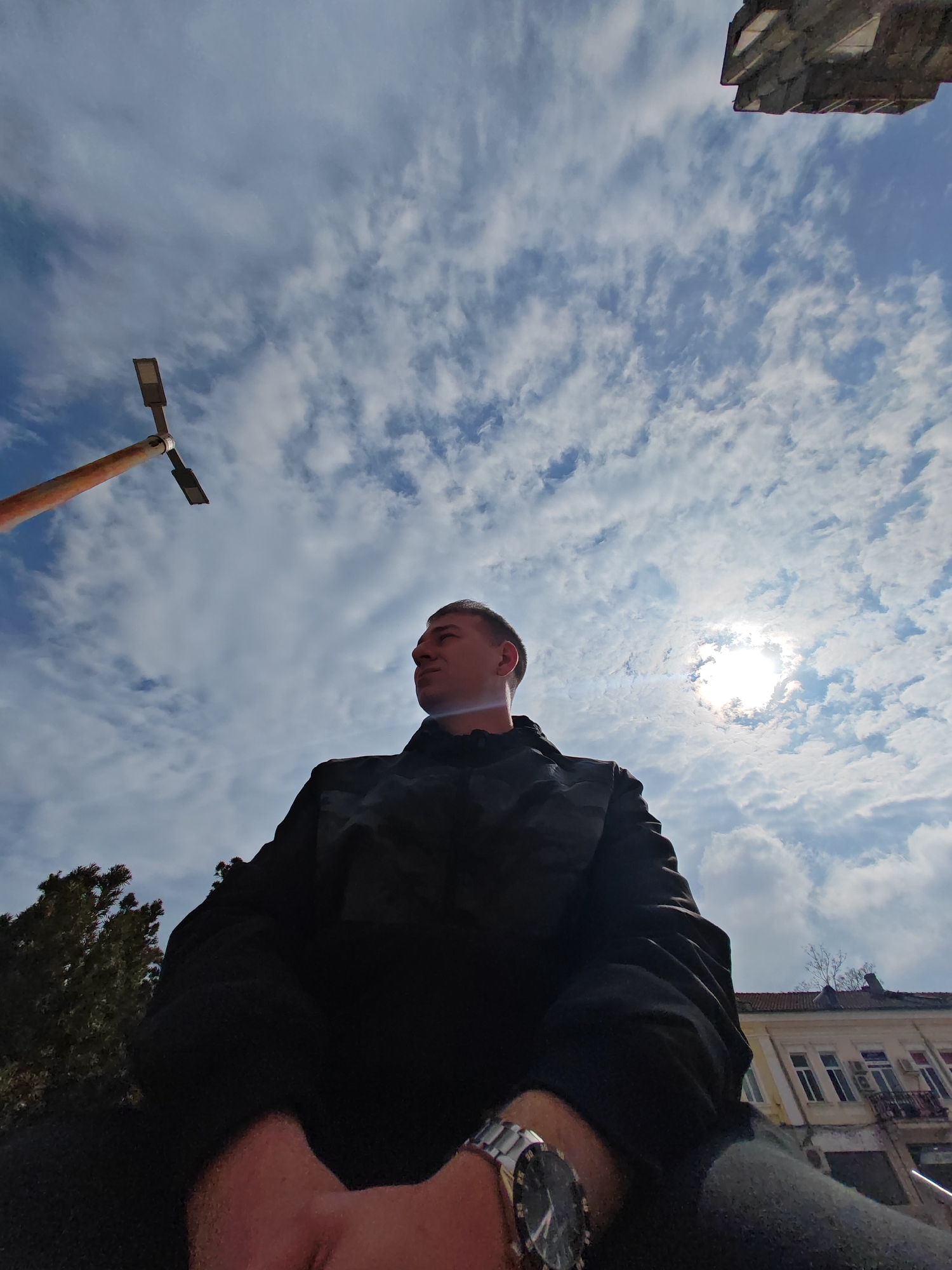 A low-angle, wide-shot photograph captures a young adult male from the chest up, positioned slightly left of center, looking upwards and towards the right. He wears a dark, possibly camouflage-patterned, jacket and a silver-toned wristwatch with a dark face on his left wrist. The sky, filled with scattered white cumulus clouds against a bright blue background, dominates the upper two-thirds of the image. The sun, intensely bright and partially obscured by clouds, generates a distinct lens flare in the upper right quadrant. To the far left, a segment of a rusty-orange streetlamp pole with two rectangular light fixtures extends into the frame. In the lower left corner, dark green foliage from a tree is visible. To the right, behind the subject, a multi-story building with a light-colored facade, multiple windows, dark rooflines, and metal balconies with attached air conditioning units is partially observed. A grey, angular architectural element is present at the extreme top right. The scene is outdoors, under direct sunlight. This depiction is of a location within the city of Shumen, Bulgaria.