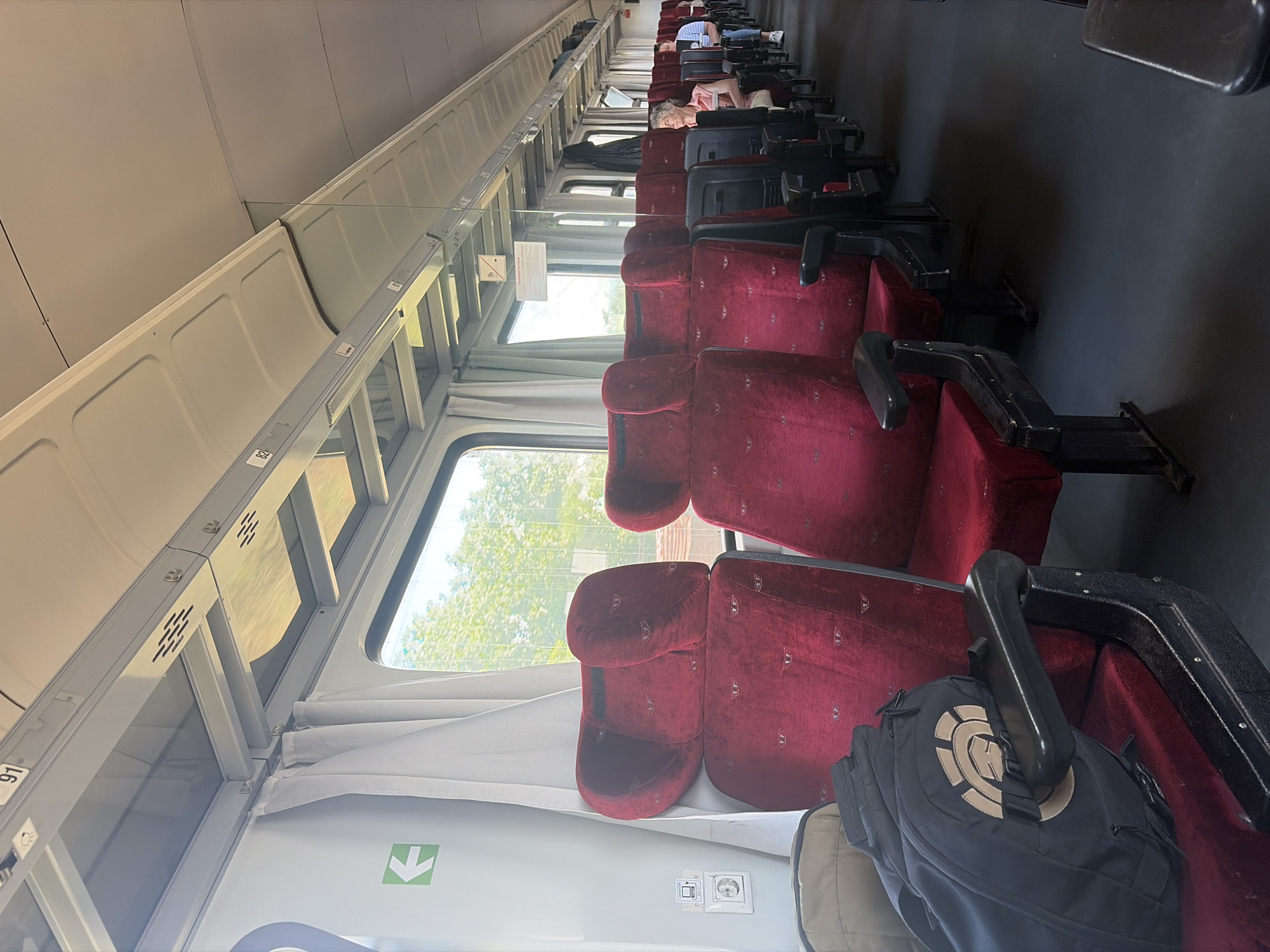 This is a photograph taken inside a train carriage, looking forward along the aisle. The seats are upholstered in a deep red velvet fabric with a subtle pattern. The carriage appears to be mostly empty, with a few passengers visible in the distance. The lighting is bright, suggesting it is daytime. The overall impression is one of a well-maintained, albeit perhaps older, train interior. The location in the city of Cherna Gora, Bulgaria, is not discernible from the image itself.