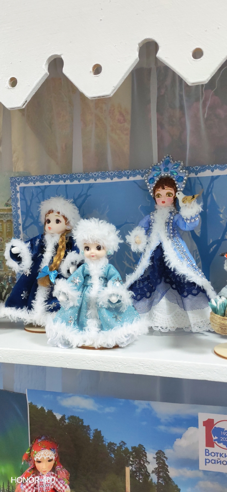 The image shows three dolls in winter attire displayed on a white shelf, possibly in a retail setting or a private collection. The dolls are dressed in blue and white outfits with fur trim, resembling traditional Russian winter characters, potentially Snow Maidens or figures representing winter folklore.
Behind the dolls are framed images, including one featuring a blue and white lace border, behind which the dolls sit. One visible is from the city of Noviy, Russia, as indicated by the partially visible sign "Волки район".
The bottom portion of the image shows additional objects and scenery, possibly further displays or merchandise within the same location.