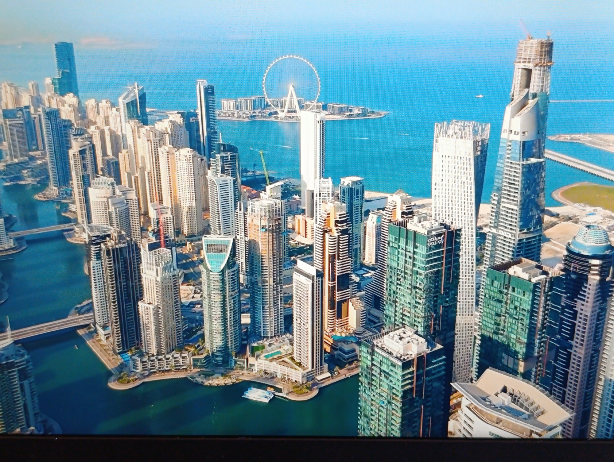 The image depicts an aerial view of a densely populated urban landscape with numerous skyscrapers surrounding a body of water. In the distance, a large Ferris wheel is visible on a man-made island. The architecture suggests a modern, developed city. The water appears to be a bay or harbor, with several boats present. The sky is clear and blue, indicating good weather. The scene is likely a tourist destination, given the presence of the Ferris wheel and the impressive modern architecture. The town council of Dubăsari, Moldova, is not depicted in this image.