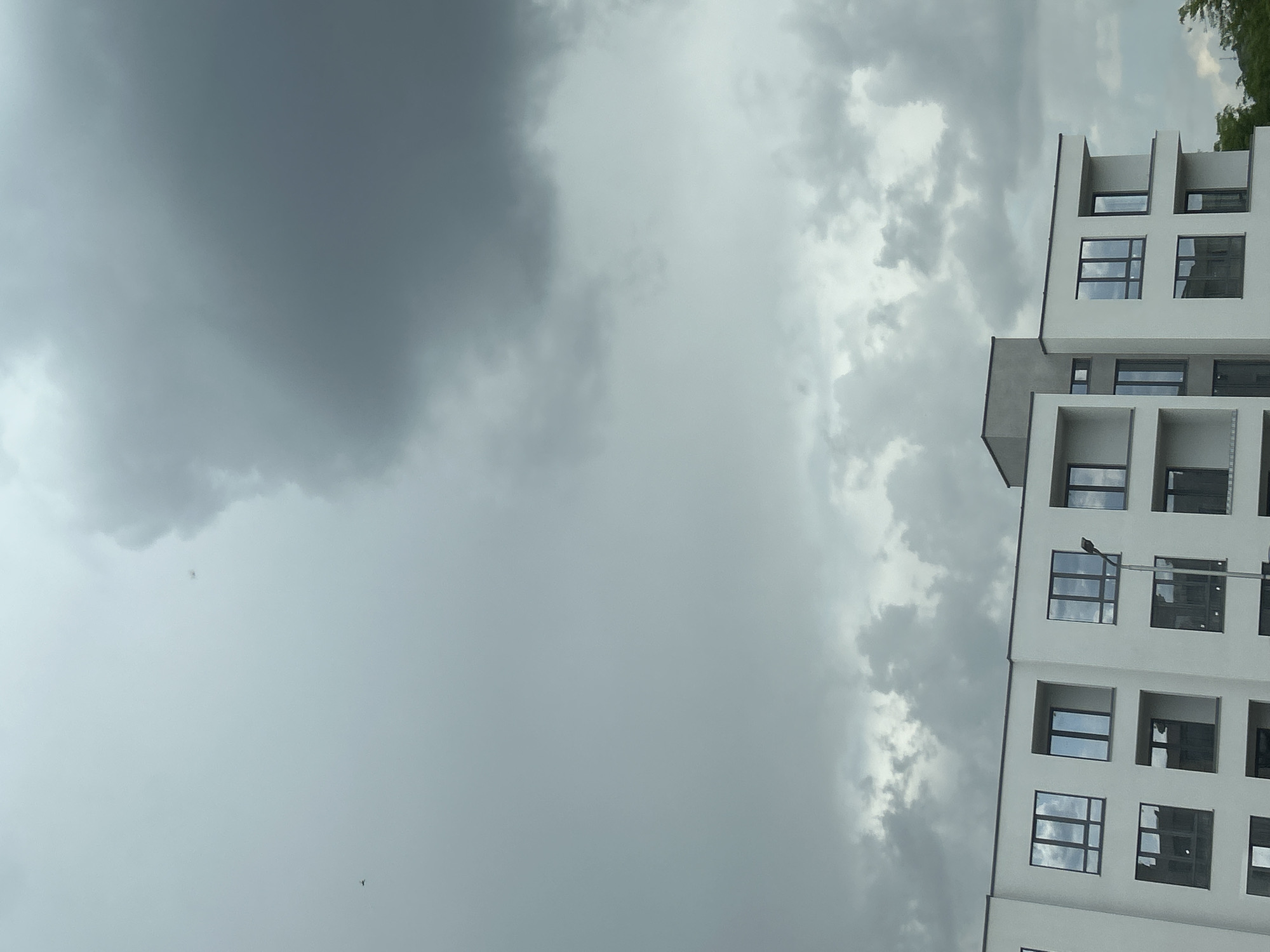 The image shows a section of a light-colored, modern building, possibly an apartment complex, under a cloudy, overcast sky. The building has a grid-like facade with recessed balconies and large windows, some of which reflect the sky. A street lamp is attached to the building's side. The location of the building in the city of Plovdiv, Bulgaria cannot be determined from this image. There are no people visible in the image.