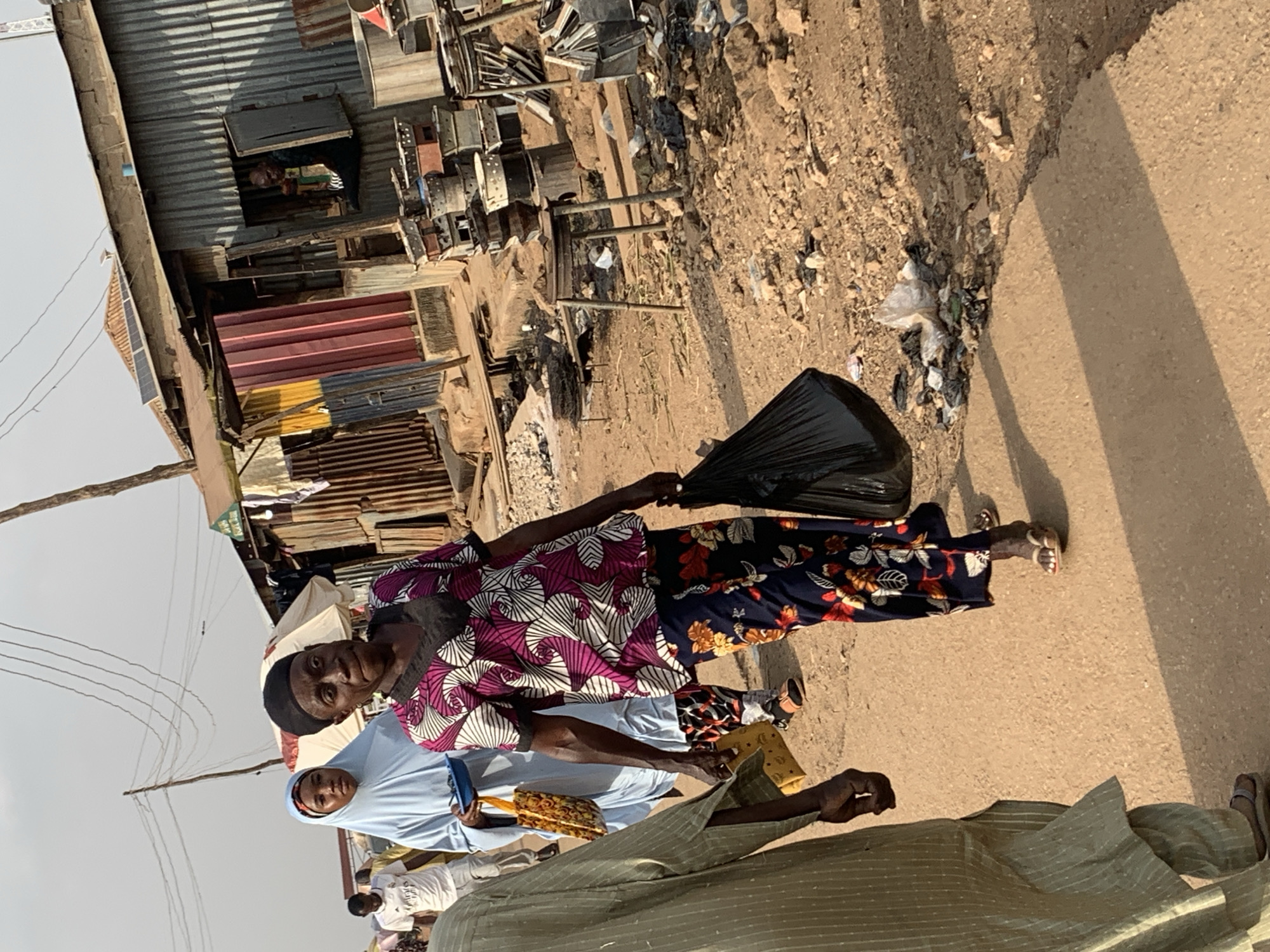 The image shows a street scene in Kafanchan B, Nigeria. An older woman in a black head covering, a pink and white patterned top, and floral-patterned pants is holding a black plastic bag. Beside her is a person wearing a light blue head covering and a light blue garment. A person in a green striped garment is also visible. They appear to be standing on a paved road. In the background, there are buildings constructed of corrugated metal and other materials, with some items stored on rooftops. There appears to be a pile of trash, and scattered trash on the side of the road. A person is visible inside one of the buildings.