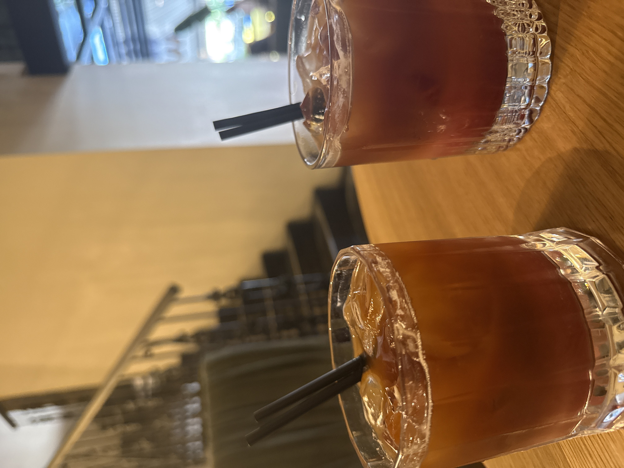 Two iced beverages with dark straws are positioned on a wooden table. The drink on the left is a deep amber color, while the drink on the right is a reddish-brown. In the background, a dimly lit staircase with a railing is visible, and further in the distance, a blurred view of a brightly lit interior space with neon lights can be seen. The scene appears to be indoors, possibly in a restaurant or bar.