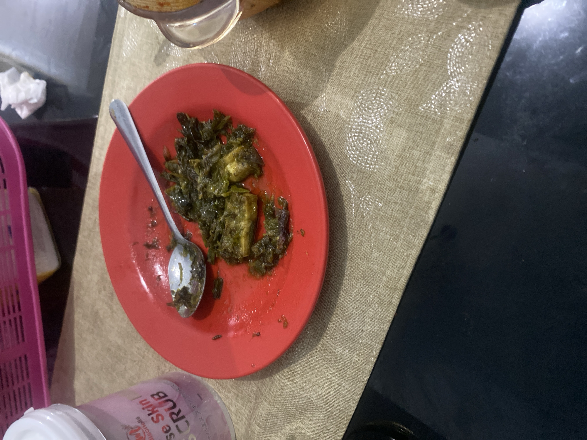 The image depicts a close-up view of a red plate containing a small portion of a green, leafy dish, possibly a vegetable stir-fry, with some chunks of what appears to be eggplant. A silver spoon rests on the plate, partially submerged in the dish. To the left of the plate, a pink plastic container with a white lid is visible, with text partially obscured but appearing to read "se Skin Scrub". The plate and container are placed on a tan-colored placemat with a textured, woven pattern. In the background, a pink plastic basket with a slatted design is present. The overall setting appears to be a domestic environment. The image does not provide any specific visual cues to determine the location within Dukuh, Indonesia.