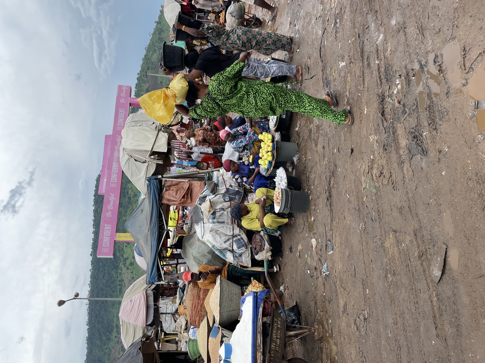 This media file captures a vibrant street market scene in Lokoja, Nigeria, likely during daylight hours given the overcast sky suggesting diffused light. The ground is a mix of mud and debris, indicating recent rain or an unpaved market area, common in many parts of Nigeria.
The scene is populated by several individuals, predominantly women, engaged in selling and managing their market stalls. They are dressed in colorful traditional attire, reflecting the local culture. One woman in the foreground wears a striking green patterned outfit and a matching headscarf, carrying a yellow bag on her head. Another woman in yellow is seated near a basket of produce, possibly lemons, and a large bucket. Several other women are visible behind them, some tending to their goods, others seated. Two men are also present, one wearing a black t-shirt and another in a camouflage-patterned outfit, both carrying black buckets.
The market stalls are constructed with various materials, including tents and tarpaulins, displaying a variety of goods which are difficult to discern in detail but appear to include foodstuffs, textiles, and possibly household items. In the background, large pink banners with white text are prominently displayed. The text "BE CONFIDENT Be Beautiful" is legible on these banners, suggesting a promotional or motivational message for the market or the community. The surrounding landscape shows a lush green hillside under a cloudy sky. The overall impression is one of a bustling local marketplace with people actively involved in commerce amidst a natural, albeit somewhat unkempt, environment.