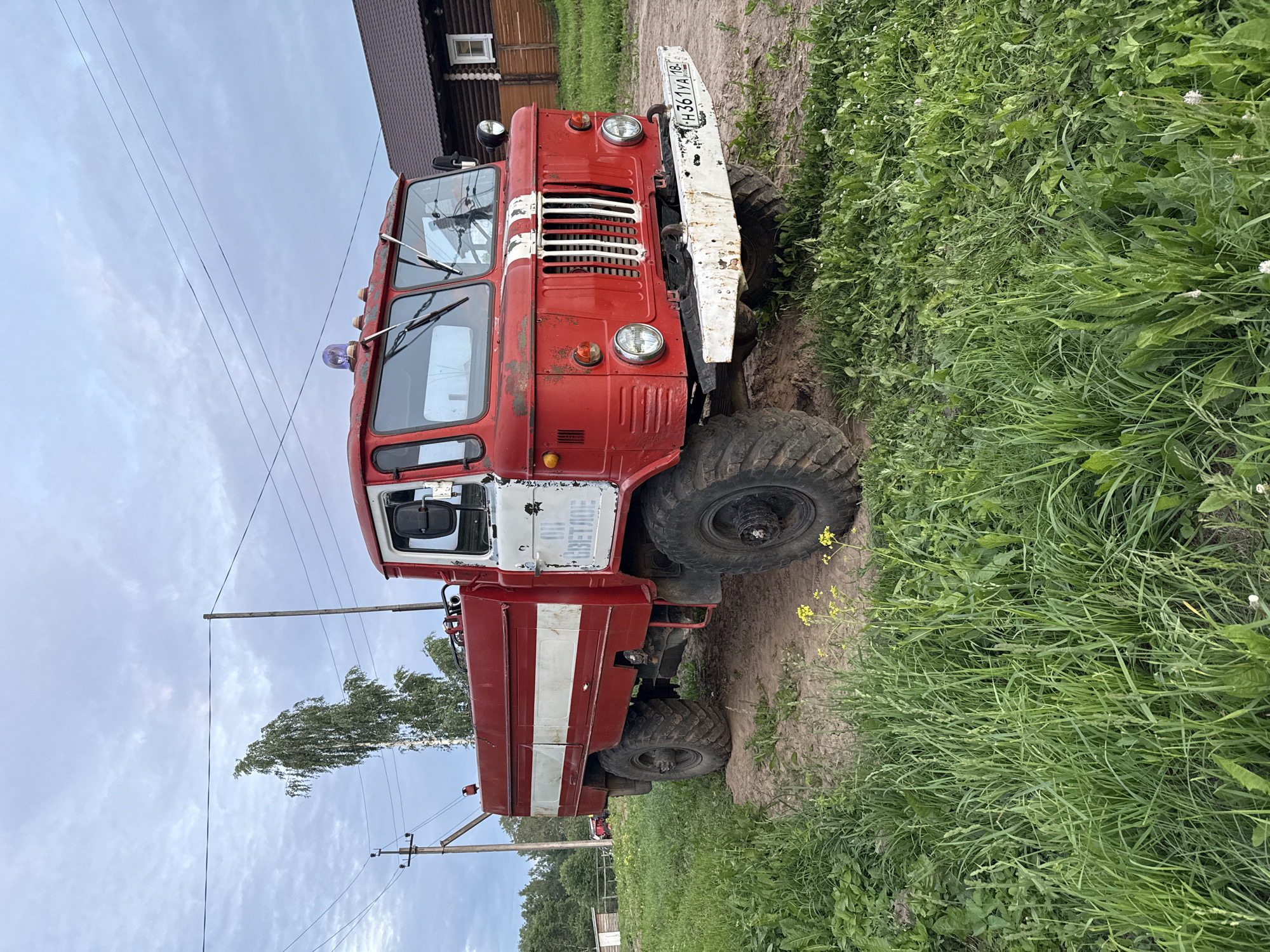 The image depicts a red fire truck with white stripes parked on a dirt path in a grassy area. The fire truck is the primary subject, positioned centrally and facing slightly to the right. It has large, rugged tires, a typical fire truck cab with visible windshield wipers and a blue flashing light on top. Text is visible on the side of the truck, appearing to be in Cyrillic script. The license plate on the front bumper is also visible and readable.
In the background, there is a wooden house with a dark roof to the right of the fire truck. To the left and behind the truck, a tall, slender tree with light-colored bark is prominent, with other trees and foliage forming a dense green backdrop. Electrical poles and wires are visible in the sky, suggesting a rural or semi-rural setting. The ground is a mix of dirt and tall green grass, with some yellow wildflowers growing in the foreground. The sky is overcast, with cloudy conditions. The scene appears to be outdoors, in what is likely a village or countryside setting. No people are present in the image. The overall impression is one of a still, quiet moment, possibly in a village like Кукуи, Russia, given the context of the provided information.