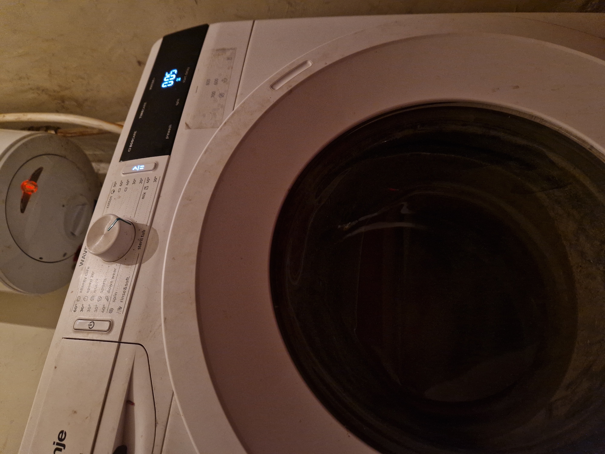 The image shows a close-up, angled view of a white washing machine in what appears to be a domestic setting. The machine has a control panel with various wash program options and a digital display showing "0:05", presumably indicating remaining time. The drum of the washing machine is visible and appears dark. The overall impression is of a functional appliance in use or recently used, located in Raduvene, Bulgaria. The environment is dimly lit, and some dust is visible on the machine's surface.