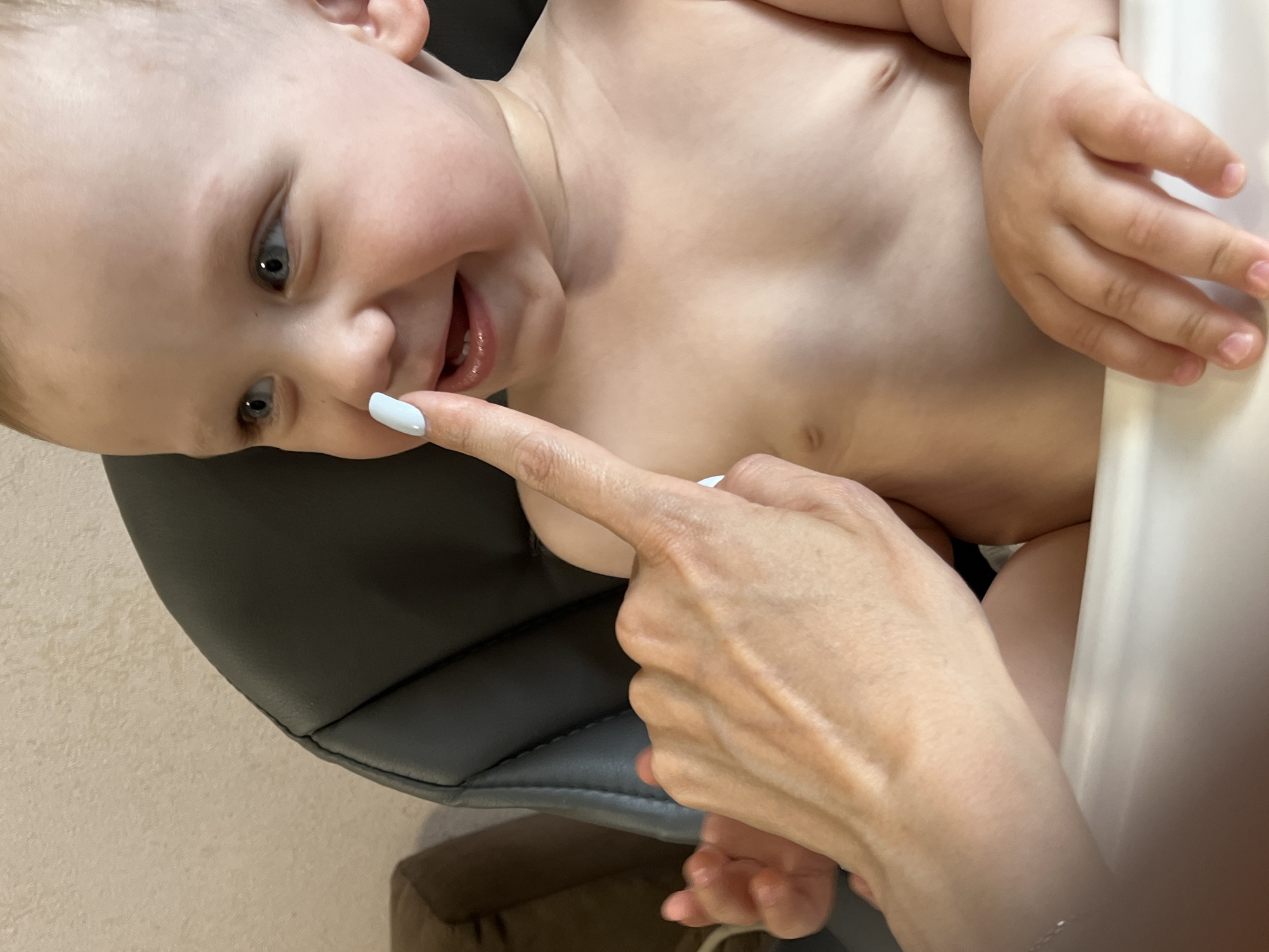 An overhead shot captures a close-up of a baby's torso and face, seated in a high chair. A woman's hand with light blue painted nails is touching the baby's nose. The baby has blue eyes and is smiling. The background is a wall and part of a chair. The image was taken in Nizhny Novgorod, Russia.