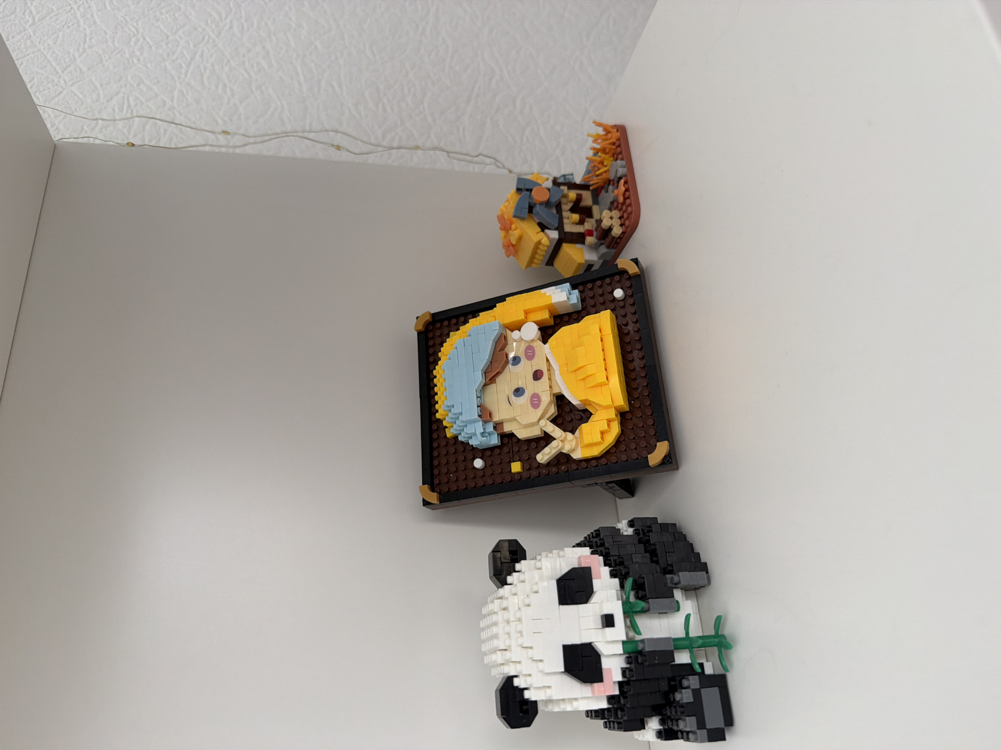 Three Lego-style building block models are displayed on a white shelf. On the left is a panda with bamboo. In the center is a framed mosaic portrait of a person wearing a yellow shirt and a blue head covering, with a pearl earring. On the right is a small building with a brown base and orange flames, resembling a fire pit or a small structure. The background is a white wall with a textured pattern. A string of small, yellow fairy lights hangs vertically on the right side of the frame. The location is not identifiable from the image.