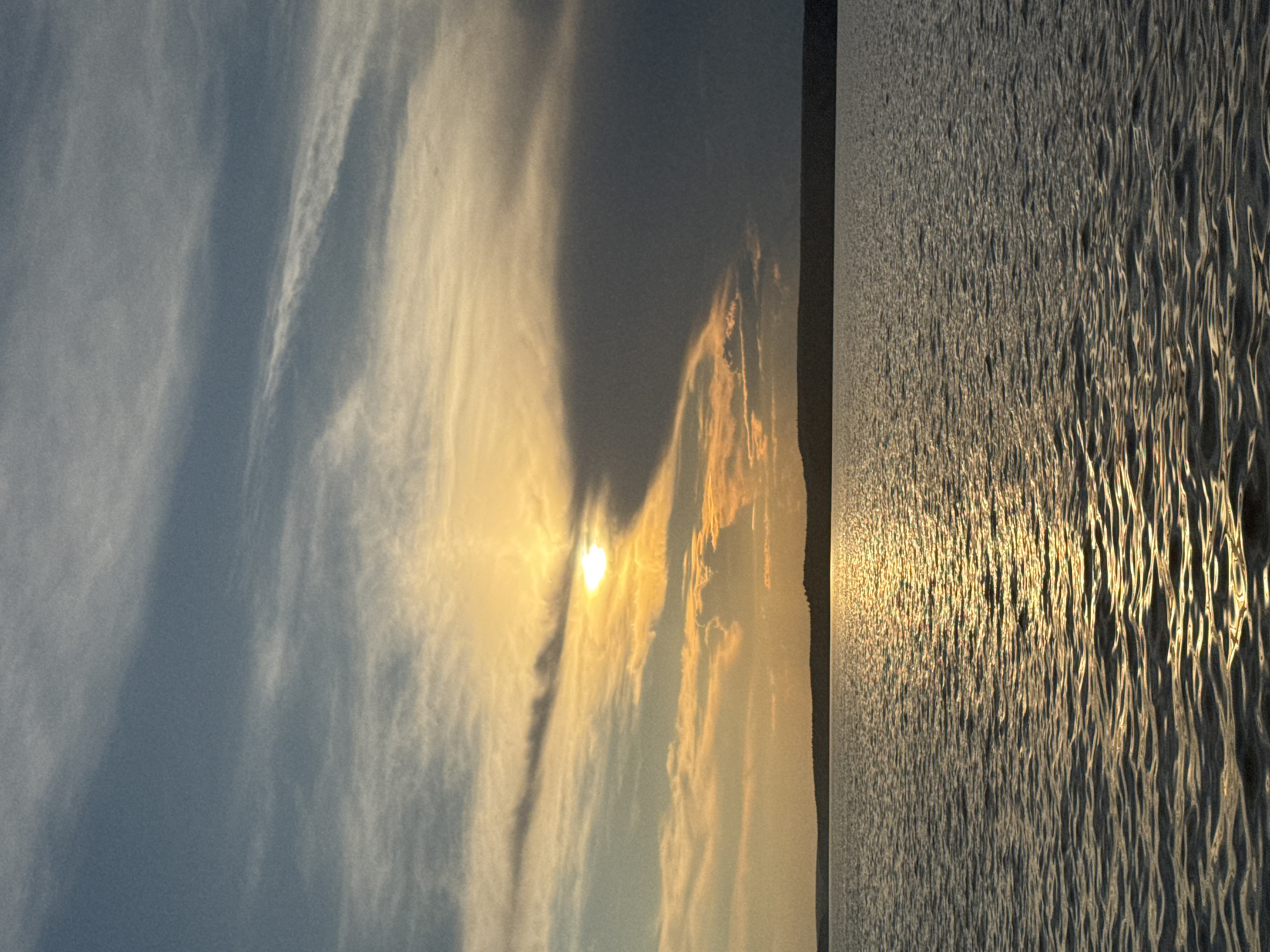 The image depicts a serene sunset over a body of water, likely a lake or large river, in Votkinsk, Russia. The sun is partially obscured by dark, dramatic clouds on the right side of the frame, while streaks of orange and yellow light emanate from behind them, illuminating the sky and reflecting brightly on the water's surface. The water itself is calm, with gentle ripples creating a textured pattern. A dark, silhouetted treeline or hillside forms a horizontal line along the horizon, separating the water from the sky. The overall atmosphere is peaceful and evocative of the end of a day in Votkinsk. There are no people or specific man-made objects clearly visible in the frame.