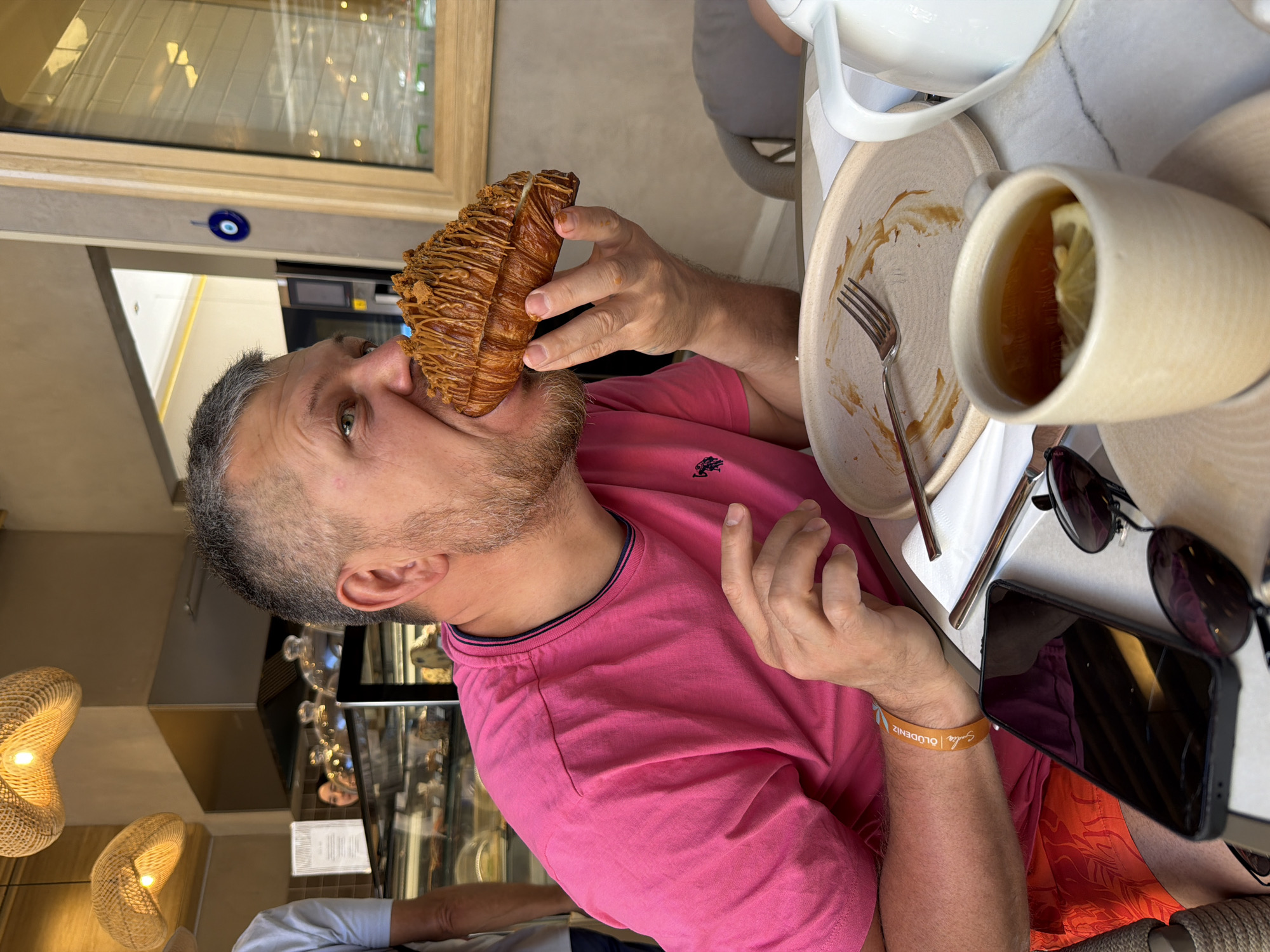The image depicts a man actively eating a large, golden-brown croissant, which is elaborately drizzled with a caramel or similar sweet sauce. The man, with short greying hair and a beard, wears a bright pink t-shirt with a small dark logo on the chest. He holds the croissant to his mouth, with his eyes wide, suggesting he is either in the process of taking a bite or playfully posing for the camera.
He is seated at a light-colored table, possibly marble. On the table to his right, a ceramic plate shows remnants of food and holds a silver fork. Next to it, a matching ceramic mug contains tea with a lemon slice floating in it, with a white teapot partially visible. A pair of dark sunglasses and a black smartphone with a cracked screen are also on the table. On the man's left wrist, an orange wristband is visible, bearing the text "ÖLÜDENİZ SUNRISE" or "SUNLIGHT."
The background indicates an indoor cafe or bakery setting during the daytime, characterized by bright light. A large window with a wooden frame shows reflections of the interior. A blue evil eye (Nazar Boncuğu) ornament is affixed near the window, a cultural symbol common in Turkey. Behind the man, display cases filled with various pastries and items are visible. Two woven pendant lights with a warm glow hang from the ceiling. Another person, partially seen in a white shirt, is seated in the background. The "ÖLÜDENİZ" wristband strongly suggests the location is Fethiye, Turkey, aligning with the provided context.