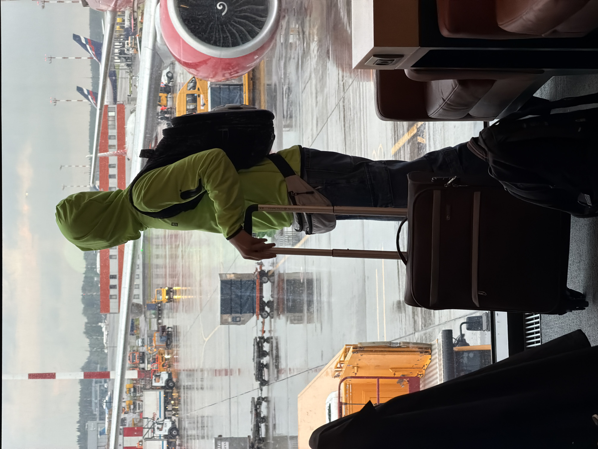 A person wearing a bright green hooded jacket stands by a large window at an airport, looking out at the tarmac. The person has a black backpack slung over one shoulder and a smaller bag strapped around their waist. In front of them is a dark brown suitcase with wheels and a handle extended. The tarmac is wet, indicating it has been raining, and several vehicles, including trucks and a yellow van, are visible. Airplanes are parked in the distance, and part of a red and white airplane engine is in the foreground on the right. The scene appears to be from Sheremetyevo International Airport in Khimki, Russia, given the presence of Aeroflot aircraft and airport infrastructure typical of that location.