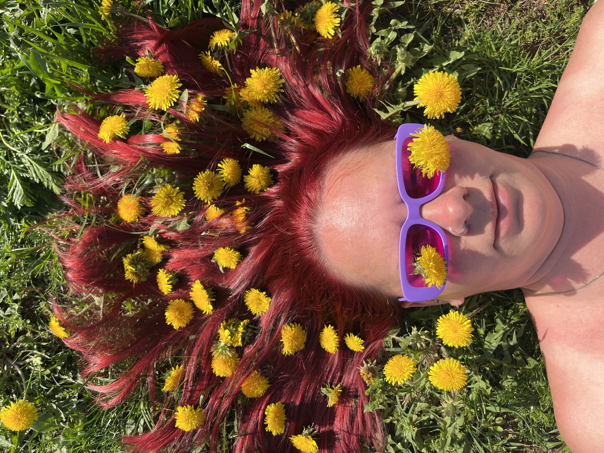 A woman lies in a grassy field. She has red hair and is wearing purple sunglasses. Her hair and sunglasses are adorned with yellow dandelions. The photo was taken in Chaikovsky, Russia.
