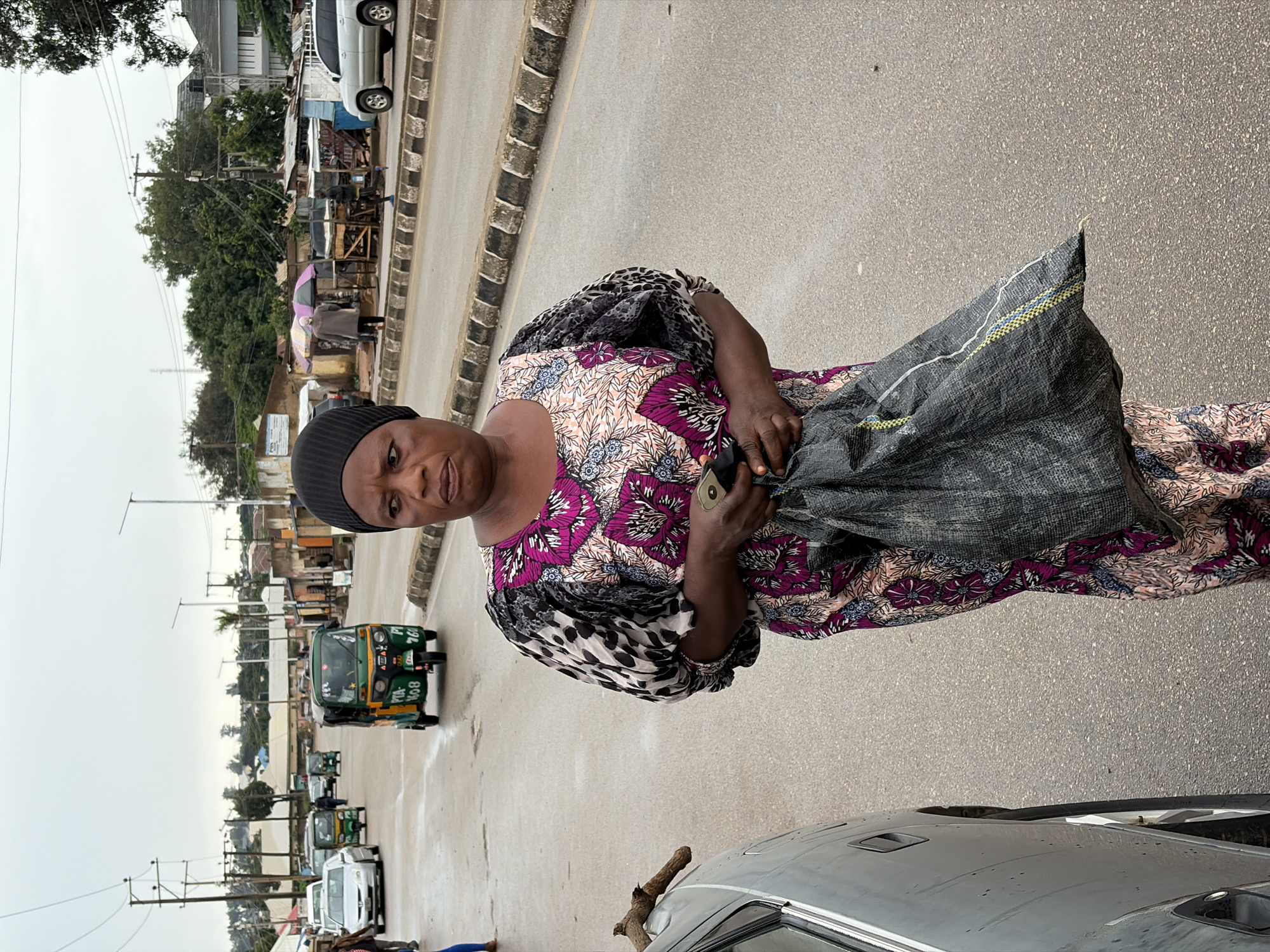 A woman wearing a black head covering and a colorful patterned dress walks on a wet, asphalt road in Jos, Nigeria. She carries a large, dark gray bag. The road is lined with a curb and a sidewalk on the right, where several stalls with awnings are visible. In the background, various vehicles, including a green and yellow auto-rickshaw, cars, and a silver SUV, are present on the road. Buildings and trees form the backdrop under an overcast sky.
