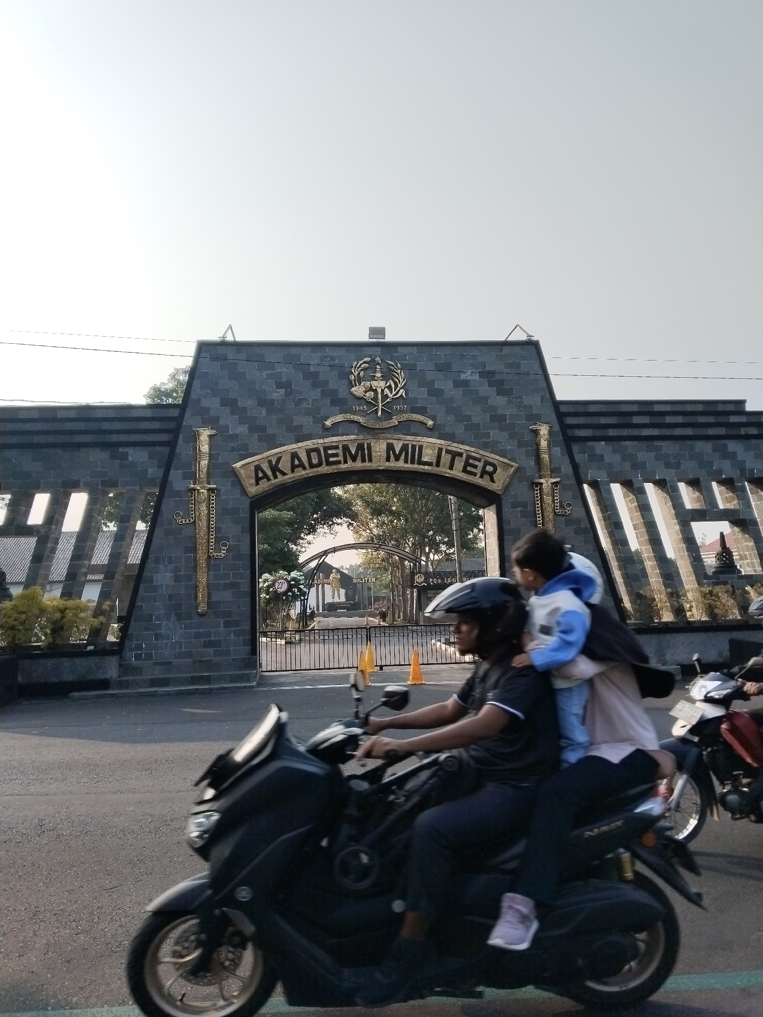 In the city of Banyurojo, Indonesia, a dark grey stone archway marks the entrance to the Akademi Militer (Military Academy). The archway is adorned with golden swords and a crest featuring the years "1945" and "1957". Below the crest, the words "AKADEMI MILITER" are inscribed in golden, arched lettering. A black motorcycle is captured in motion in the foreground, carrying three occupants. The rider, wearing a black helmet and t-shirt, is steering the motorcycle. Behind the rider, a person wearing a headscarf and a light-colored jacket is seated, holding a child dressed in a blue and white jacket. The child is facing forward, looking towards the academy entrance. The motorcycle is on a paved road, with a black metal fence and the academy grounds visible beyond. Another motorcycle is partially visible to the right. The sky is bright and overcast.