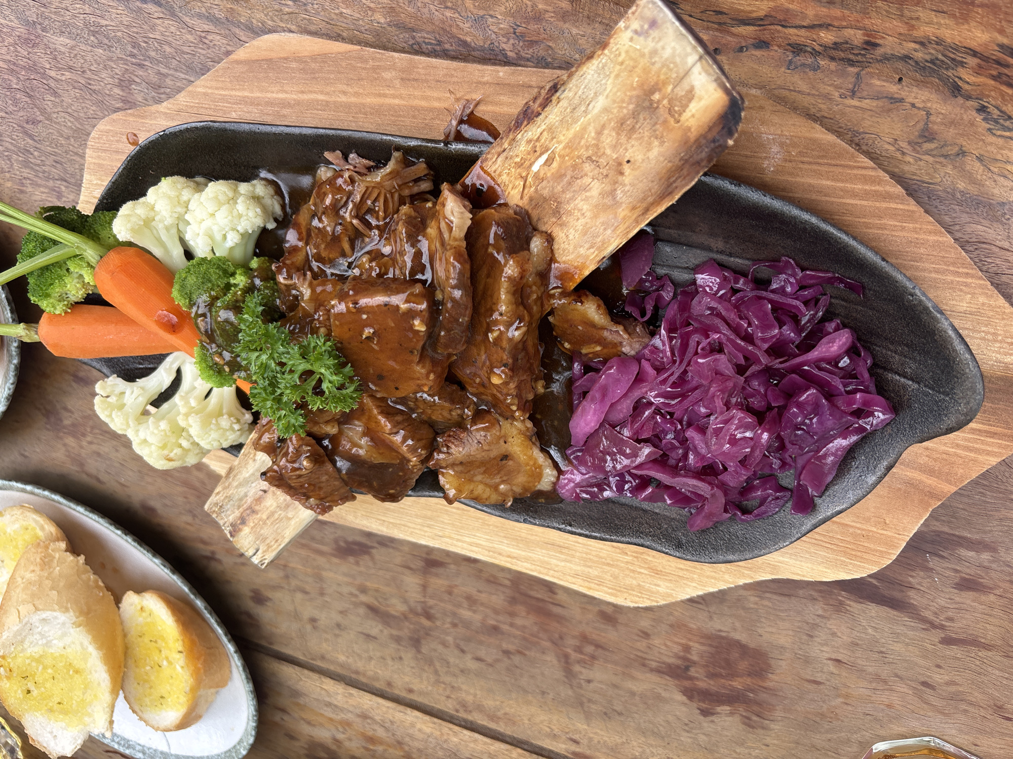 A close-up, eye-level shot depicts a dish of what appears to be short ribs with vegetables and bread, set on a wooden table in an outdoor setting. The dish is served on a dark, irregularly shaped platter placed on top of a lighter wooden board. The short ribs are drenched in a dark sauce, interspersed with two large bone sections. Accompanying the ribs are blanched cauliflower florets, raw carrots, broccoli, and cooked red cabbage. Fresh parsley provides a garnish. A small dish containing buttered bread slices is partially visible in the lower-left corner of the image. The setting suggests an outdoor dining area, potentially in the city of Da Lat, Vietnam. The wooden table and the natural light indicate an informal eating environment.