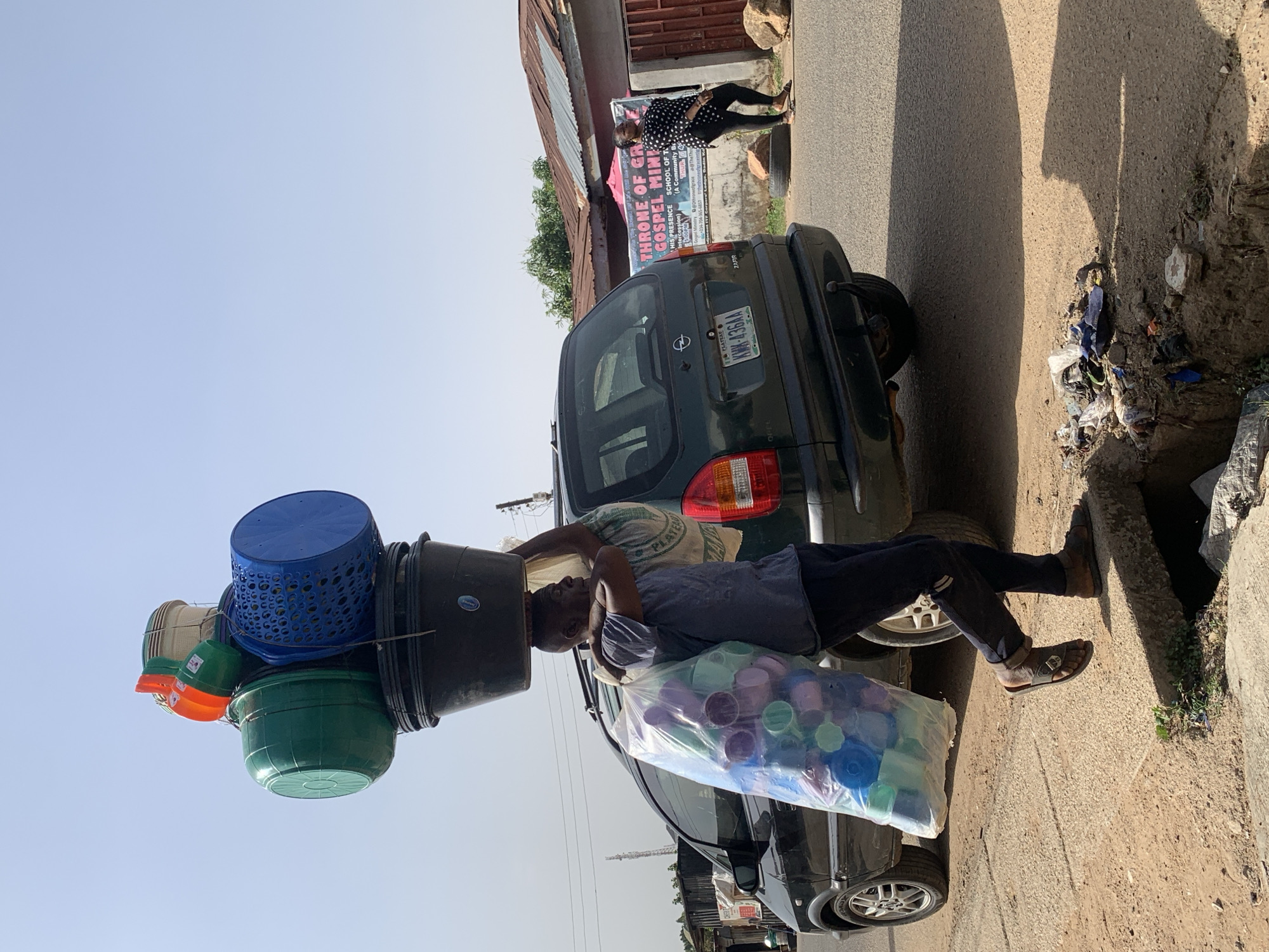 A man in Jos, Nigeria carries a stack of buckets on his head and a bag of plastic cups while standing next to a dark green Opel Zafira with the license plate KMK-436AA. The street scene includes a woman in a black and white polka dot outfit, walking near a sign for the Throne of Grace Gospel Ministry. Debris is visible along the roadside.