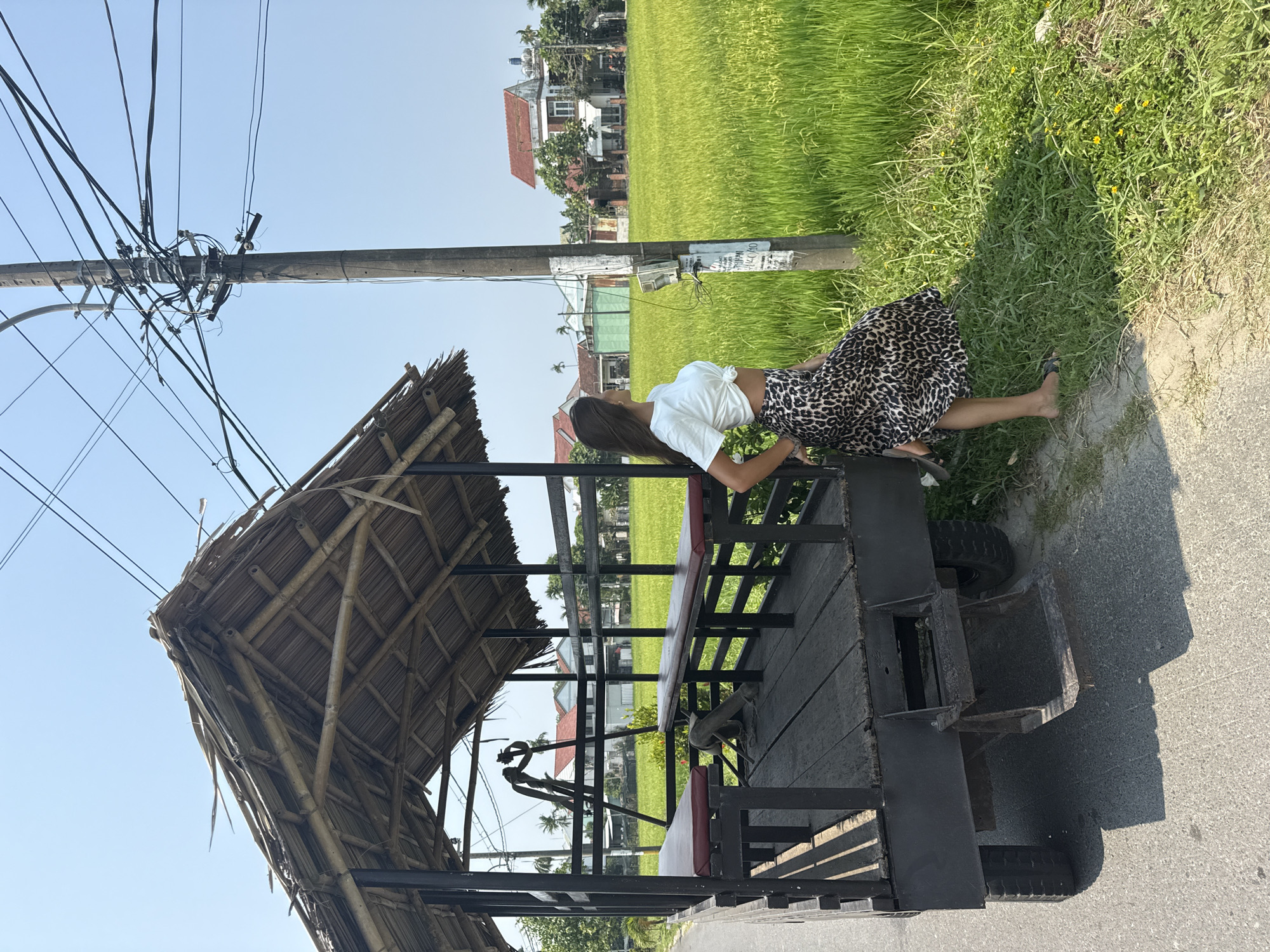 A young woman in a white tied top and leopard-print skirt leans against a black flatbed trailer with a bamboo roof, on the side of a road. The flatbed has benches inside. A telephone pole with wires stands beside the trailer. In the background, there is a green rice field, houses, and a blue sky. The location is likely in a rural area of Đà Nẵng, Vietnam, given the presence of rice fields and the style of houses. The woman is barefoot or wearing sandals.