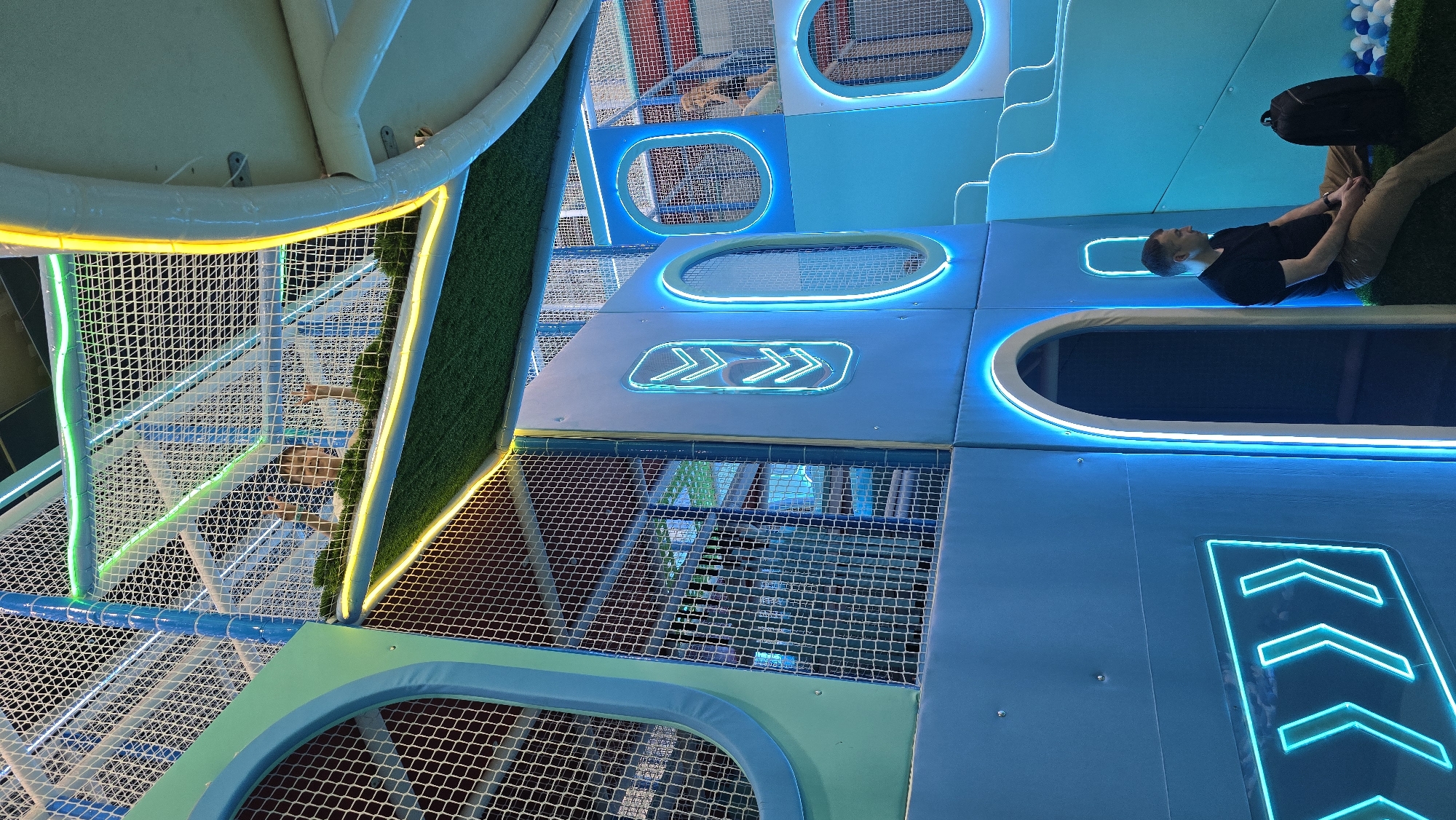 The image depicts an indoor, multi-level play structure, likely a soft play gym or adventure park, in an unknown city and state. The dominant color scheme is shades of blue and teal, accented by extensive LED strip lighting in blue, green, and yellow. White safety netting encloses various levels of the structure.
In the upper section, several children are visible through the white netting. One child, positioned centrally, is smiling and has both hands raised in a waving gesture. Portions of the upper levels feature green artificial turf. Green and yellow LED strips illuminate the edges of these elevated platforms.
The main structure consists of padded, modular panels in light blue. These panels feature circular and arched openings, all outlined with bright blue LED strips. Additionally, decorative triple chevron arrow patterns, rendered in blue LED light, are affixed to the vertical panels.
At the lower right, a man is seated on a low, turf-covered ledge. He wears a black short-sleeved shirt and light brown pants, with a black backpack resting beside him on the turf. He has short dark hair and is looking towards his right. Behind him and to his right, a shallow ball pit containing blue, white, and gray plastic balls is visible. To the left of the man, a large arched entrance, outlined with blue LED light, provides access into the structure. In the background to the right, blue multi-tiered steps or slides are apparent, along with a wall featuring red and white striped panels.