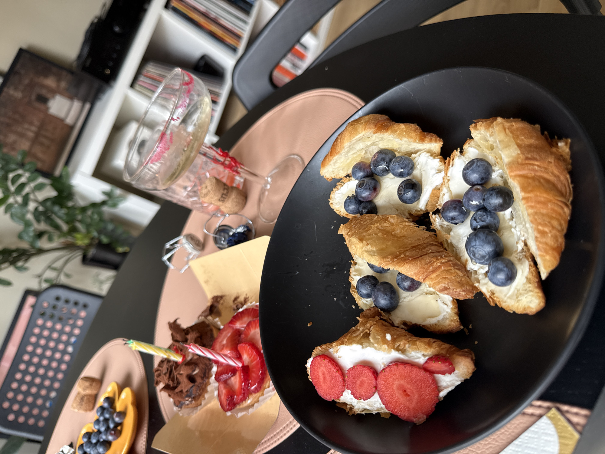 A black plate of croissant halves topped with cream and blueberries, and one with sliced strawberries, sits on a black table. Another plate nearby contains a chocolate and strawberry tart with lit birthday candles, as well as a tart with blueberries and what appears to be mango. A wine glass with lipstick marks and a cork are also on the table. The location is indeterminate, but could possibly be in Barcelona, Spain. The scene suggests a celebration, potentially a birthday. No people are visible.