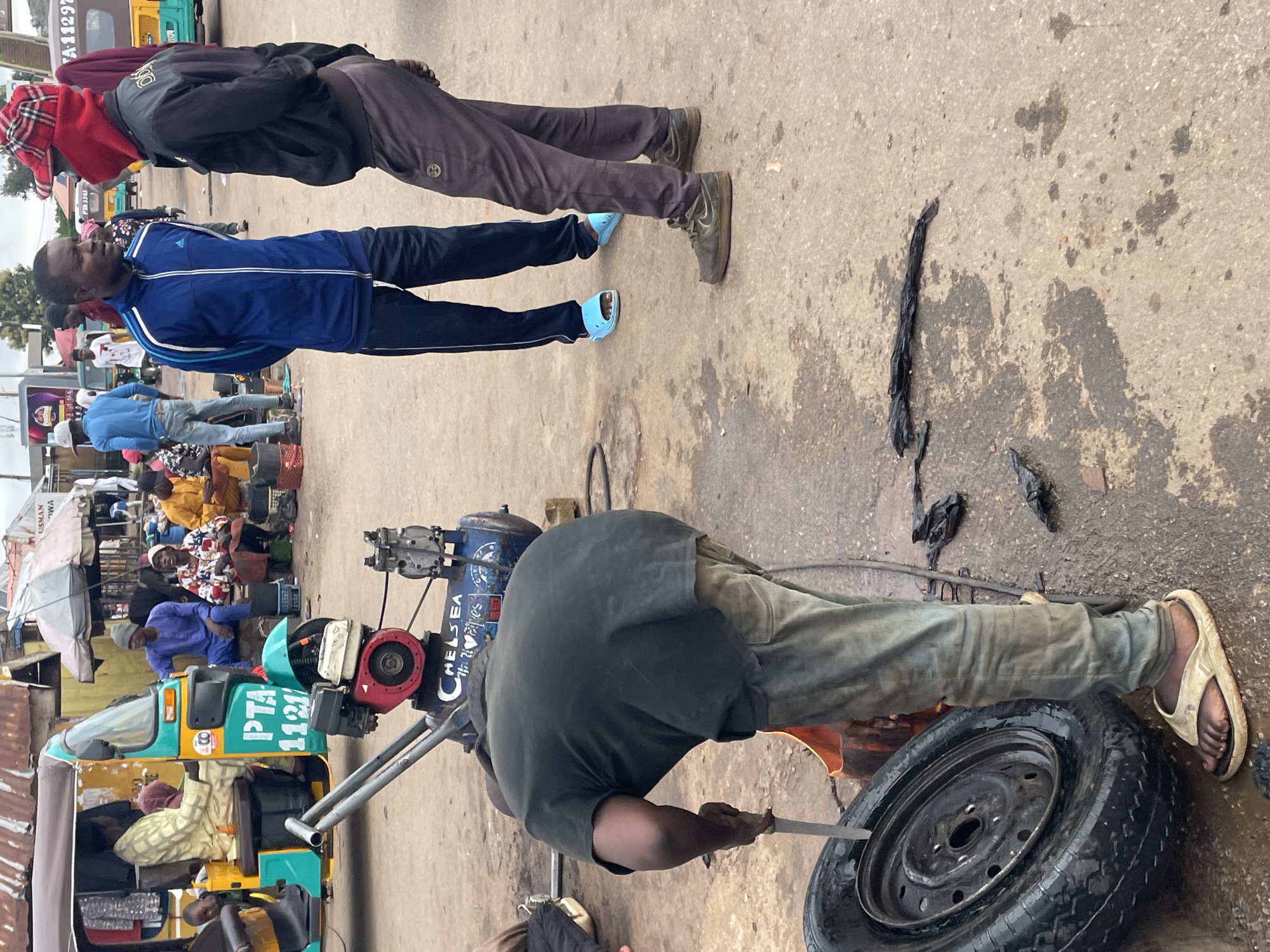 In Jos, Nigeria, on a dusty, wet street, a man in a black t-shirt and grey pants is focused on a tire repair. He is bent over, holding a knife in his right hand, and appears to be working on the tire with a tool next to it. The tire, a standard car tire, rests on the ground. Behind him, a portable air compressor, branded "Chelsea," is visible. In the background, a three-wheeled auto-rickshaw, a common mode of transport in Nigeria, is parked, with people around it, suggesting a busy marketplace or transport hub. The overall scene depicts a slice of everyday life in a Nigerian city, with a focus on informal commerce and repair work.