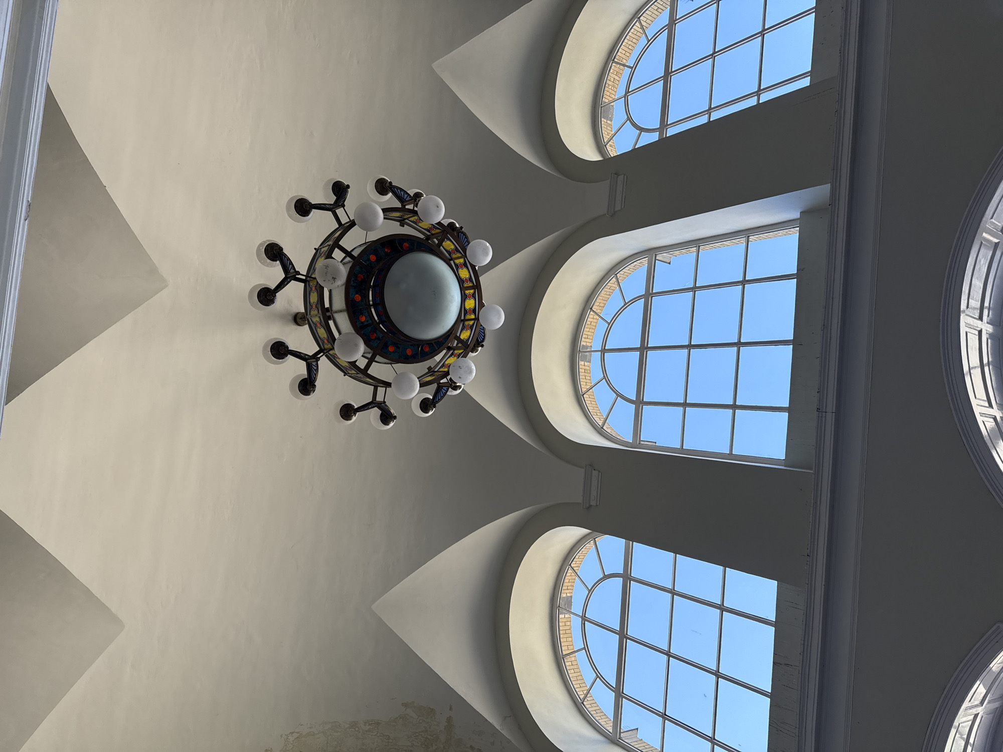 The image presents an upward perspective of a grand interior ceiling, focusing on a large, elaborate chandelier and several arched windows. A decorative chandelier with a central spherical light and numerous smaller, white globe-shaped bulbs arranged in a circular pattern hangs prominently from the ceiling. The chandelier's frame features intricate detailing with what appears to be colorful stained glass or patterned elements.
The ceiling, painted in a light, neutral tone (likely off-white or light beige), showcases architectural elements like subtle vaults and angular sections. Along the right side of the frame, three large, arched windows are visible, allowing abundant natural light to flood the space and revealing a clear, vibrant blue sky outside. These windows are divided into multiple rectangular panes by a grid of white mullions. The windows are set within deep, curved recesses in the ceiling structure, creating distinct patterns of light and shadow on the surrounding surfaces. Brickwork is faintly visible around the exterior frames of some windows.
The photograph captures a sense of architectural grandeur and the natural illumination of the space during clear daytime weather. No people or specific activities are depicted, and no text is visible within the frame. The scene suggests a public building or a large, ornate hall, consistent with a location in Szeged, Hungary.
