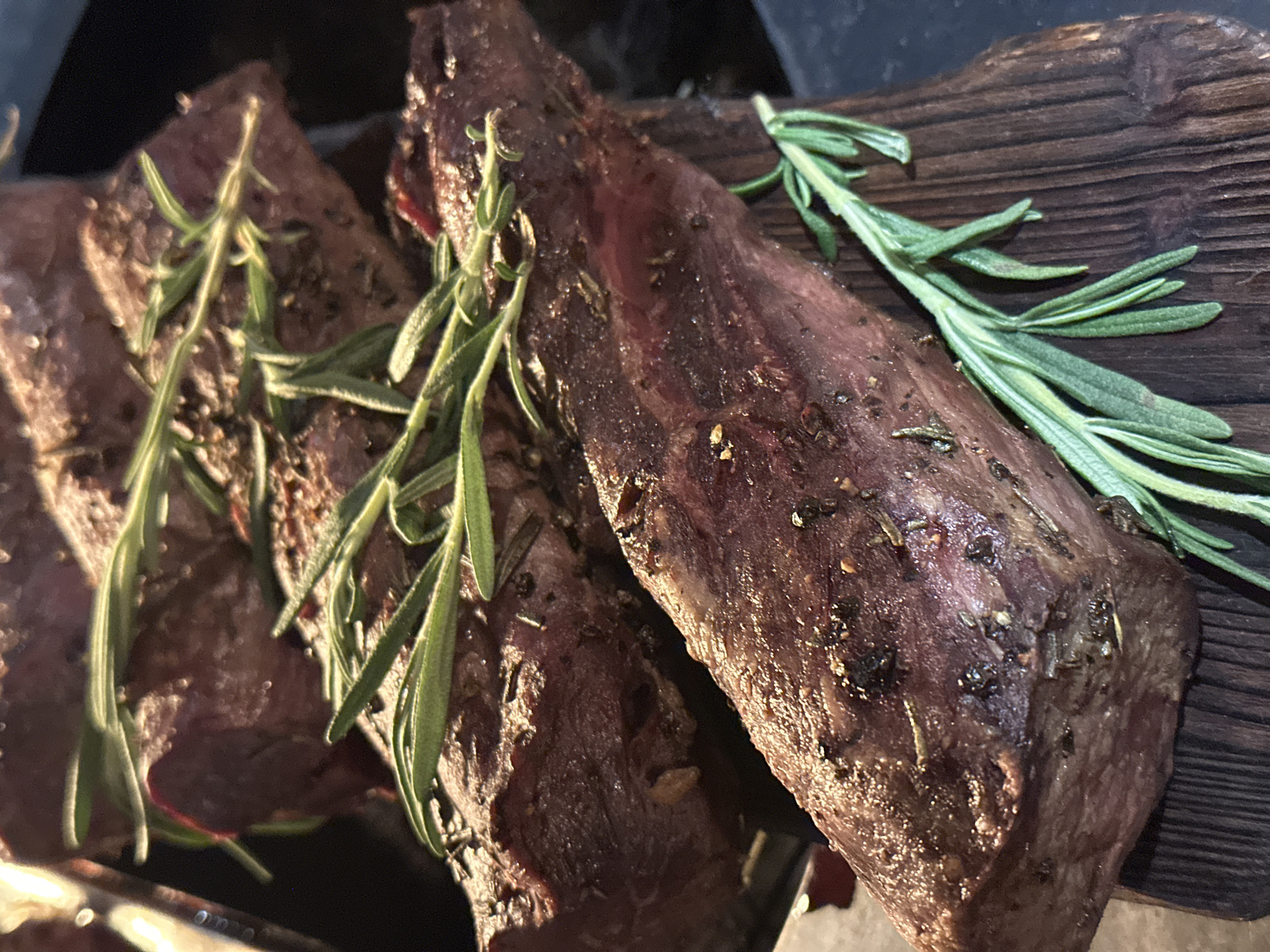The image shows three pieces of grilled meat, possibly steak, garnished with fresh rosemary sprigs, arranged on a dark wooden board. The meat is browned, suggesting it has been cooked over high heat. The rosemary appears to be freshly cut. The scene is lit with a warm light, accentuating the textures of the meat and wood. There is no indication of the location being Новоглаголево, Russia.