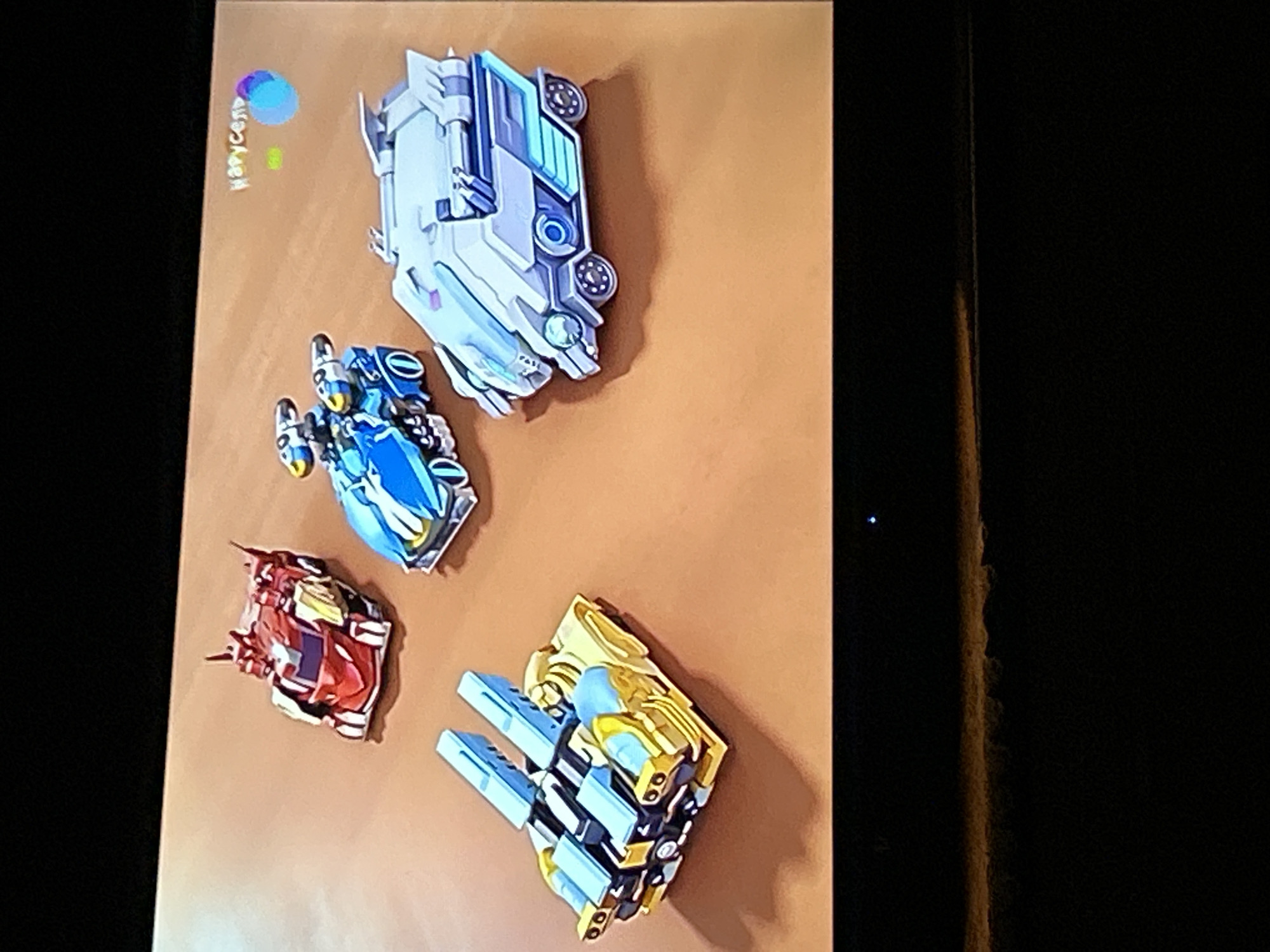 The image displays four toy vehicles on a brown surface. From left to right, top to bottom, the vehicles are: a red and silver robot-like car with pointed elements on its head and shoulders, a blue and yellow futuristic car with two pods on its back, a gold and blue vehicle with two large blue cannons mounted on top, and a white and light blue armored vehicle with a sleek, angular design. In the upper right corner, a logo with the word "Карусель" and a colorful circle is visible, which is the logo for the Russian children's television channel Karusel. The environment appears to be a studio setting or a digitally rendered scene, as suggested by the uniform brown surface and the absence of natural elements. There is no discernible indication of the city of Gelendzhik, Russia, in this image. The bottom portion of the image is dark, obscuring any potential details of the foreground or surrounding environment.