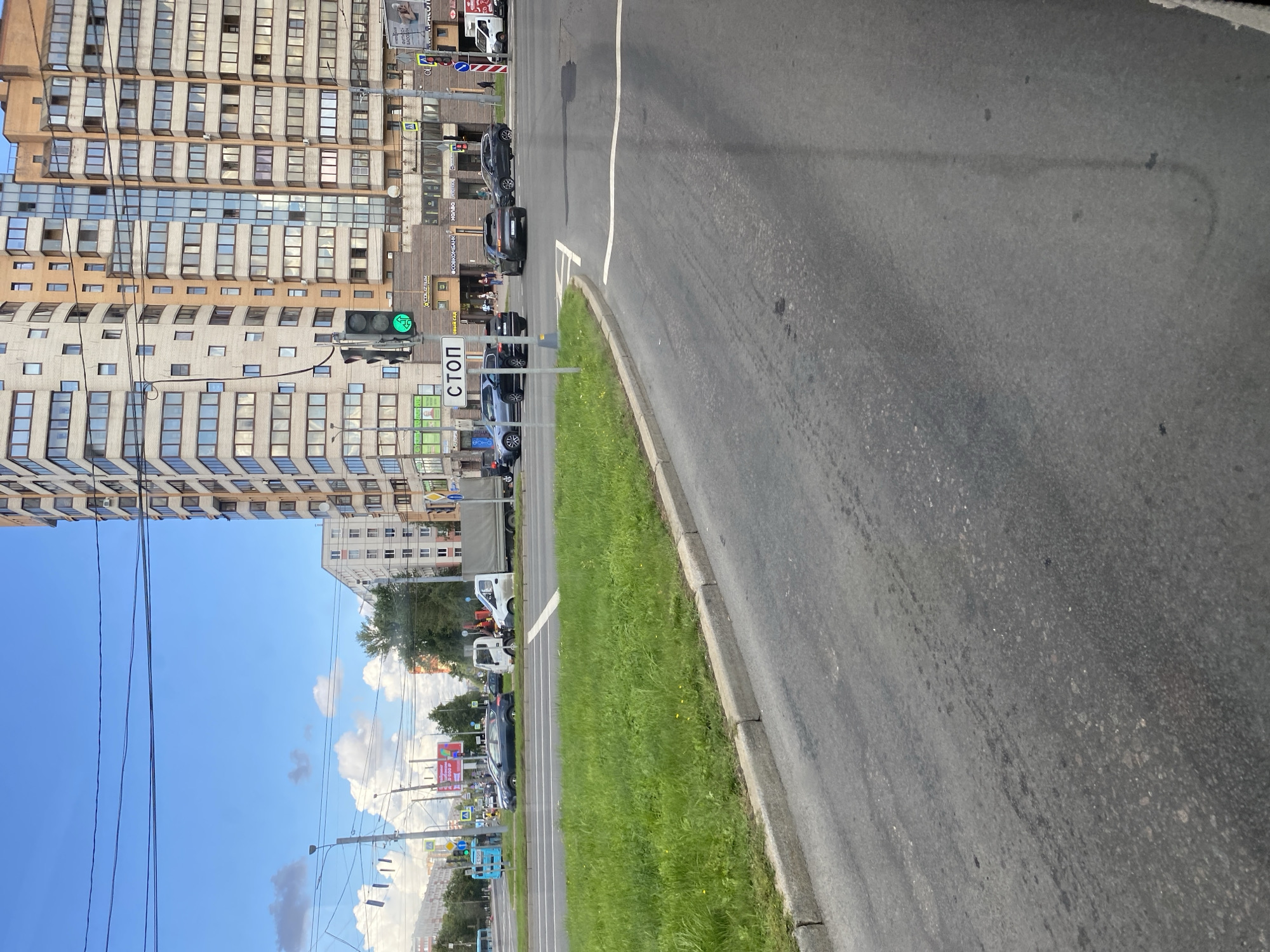 The image depicts a street scene in Saint Petersburg, Russia, on a bright, cloudy day. In the foreground, a paved road curves to the right, with a concrete curb and a patch of green grass. In the midground, several cars are visible on a multi-lane road. One car, a dark sedan, appears to be stopped at an intersection. A white truck with a gray cargo bed is parked in the right lane. Further down the road, another white truck is visible.
A large, modern apartment building with many windows dominates the background. The building is light brown and beige in color, with some glass balconies. A traffic light is visible on the left side of the image, showing a green signal. A "STOP" sign is also clearly displayed. A pedestrian can be seen walking on the sidewalk in front of the building. The sky is blue with scattered white clouds. The overall impression is that of a typical urban environment in Saint Petersburg.