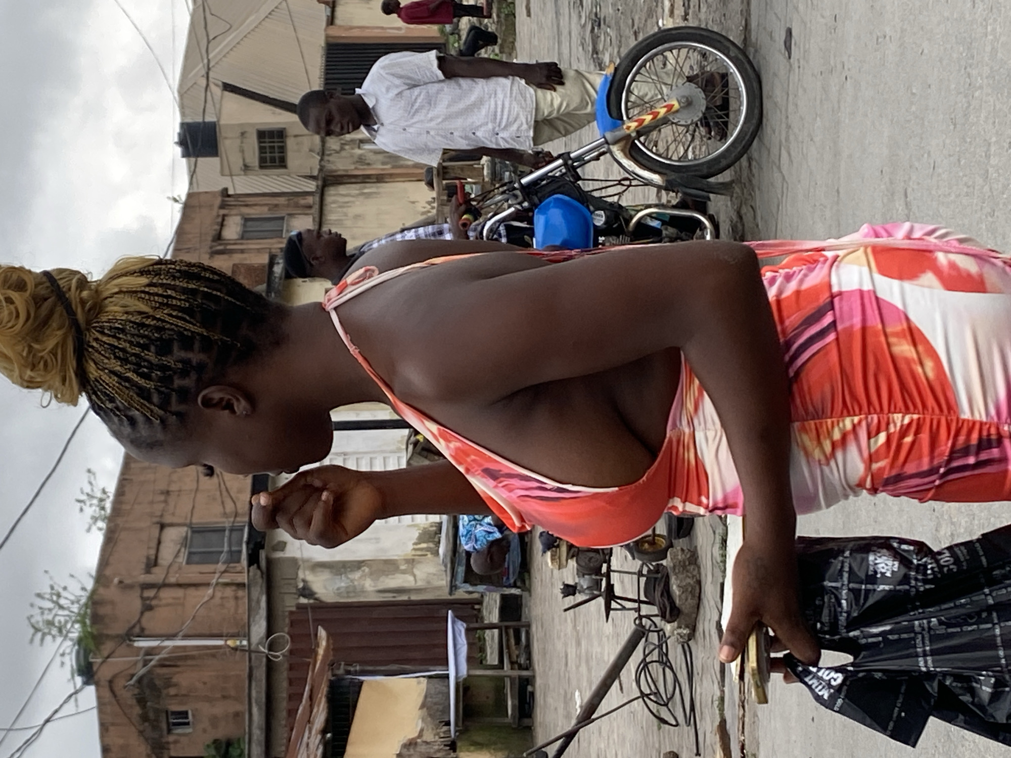 In a city environment, a woman with braided blonde hair styled in a bun stands in profile, facing left, while holding a black plastic bag and a small golden object. She wears a brightly colored, patterned dress with plunging neckline that reveals her cleavage. Her right hand is near her mouth, and her left hand holds the bag and object. In the background, several people are visible, including two men standing near a blue motorcycle. One man in a white shirt and khaki pants stands to the right, while another man in a plaid shirt stands behind him. Another man in a red shirt is further in the distance to the right. The scene includes buildings with weathered facades, electrical wires overhead, and what appears to be street equipment in the foreground. The ground is a gray, paved surface.