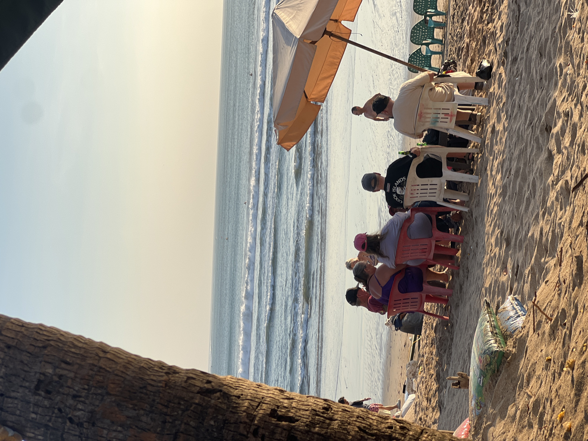 This is a vertical, eye-level, medium shot of a beach scene in Kuta, Indonesia, at sunset. In the foreground, the trunk of a palm tree is partially visible on the left. A group of people are seated on chairs on the sand, facing the ocean. Several individuals are facing away from the camera, engaged in conversation. One person is visible from the front, wearing a pink hat. A large orange beach umbrella is erected on the right side of the frame. The ocean, with gentle waves and a few surfers in the distance, stretches across the middle ground. The sky is a pale yellow and blue, indicating sunset. The sand is a warm brown, with some scattered debris and bags. The overall atmosphere is relaxed and casual.