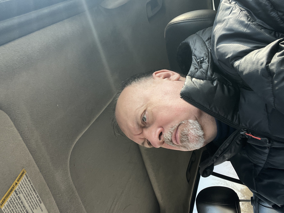 An adult male, appearing middle-aged or older with a fair complexion, balding scalp, and a grey goatee, is depicted seated inside a vehicle in Ерино, Russia. He wears a black puffy winter jacket over a dark turtleneck and is looking upward and to his left. The vehicle's interior features a light grey headliner, and a yellow warning label ("AVERTISSEMENT / WARNING") is partially visible on the sun visor. Strong sunlight illuminates the scene from the right. A dark headrest is behind the subject. The exterior visible through the window on the left shows bare tree branches against a bright sky.