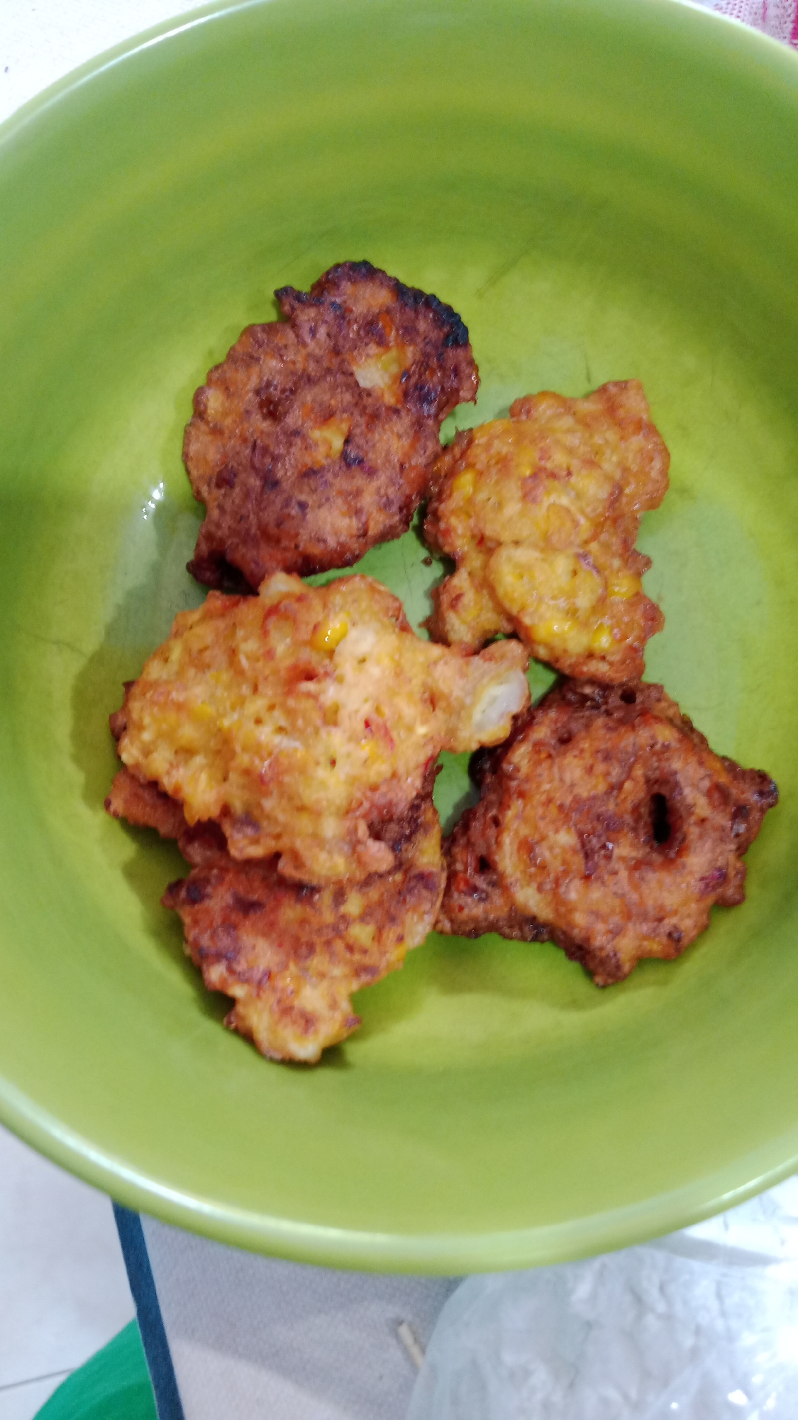 Five corn fritters are arranged in a light green ceramic bowl. The fritters are golden brown and appear to be made with corn kernels and possibly some chili peppers. The bowl is positioned on a light-colored, slightly textured surface, likely a table. The background is blurred, but suggests an indoor setting. No people are visible in the image. This image was taken in Denpasar, Indonesia, inside a residential kitchen, likely during meal preparation or serving.