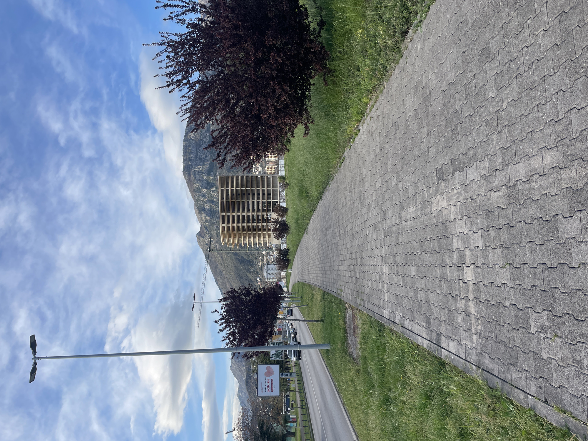 The image depicts a pedestrian walkway paved with gray interlocking bricks. The walkway is bordered by green grass on one side and a road with a sidewalk on the other. In the background, there is a mountain range partially obscured by clouds. A tall building under construction, with a skeletal frame, is visible in front of the mountain. A construction crane is also present. A road with vehicles is visible. There is a street lamp on the left and a billboard is on the side of the road. The image is likely taken in Bar, Montenegro, based on the background landscape and urban features.
